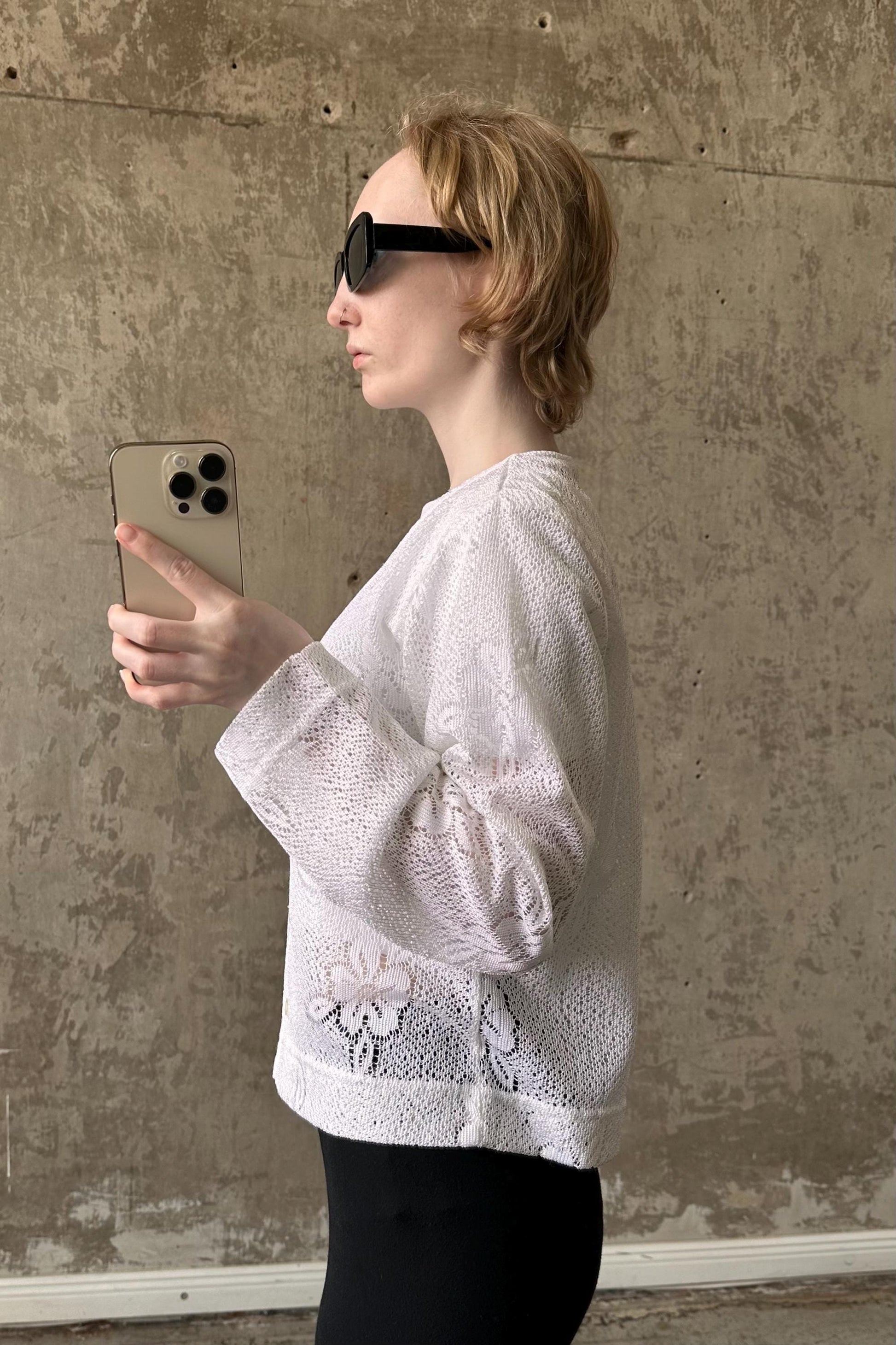 A woman wearing a white lace top and sunglasses is holding a smartphone and looking at it.
