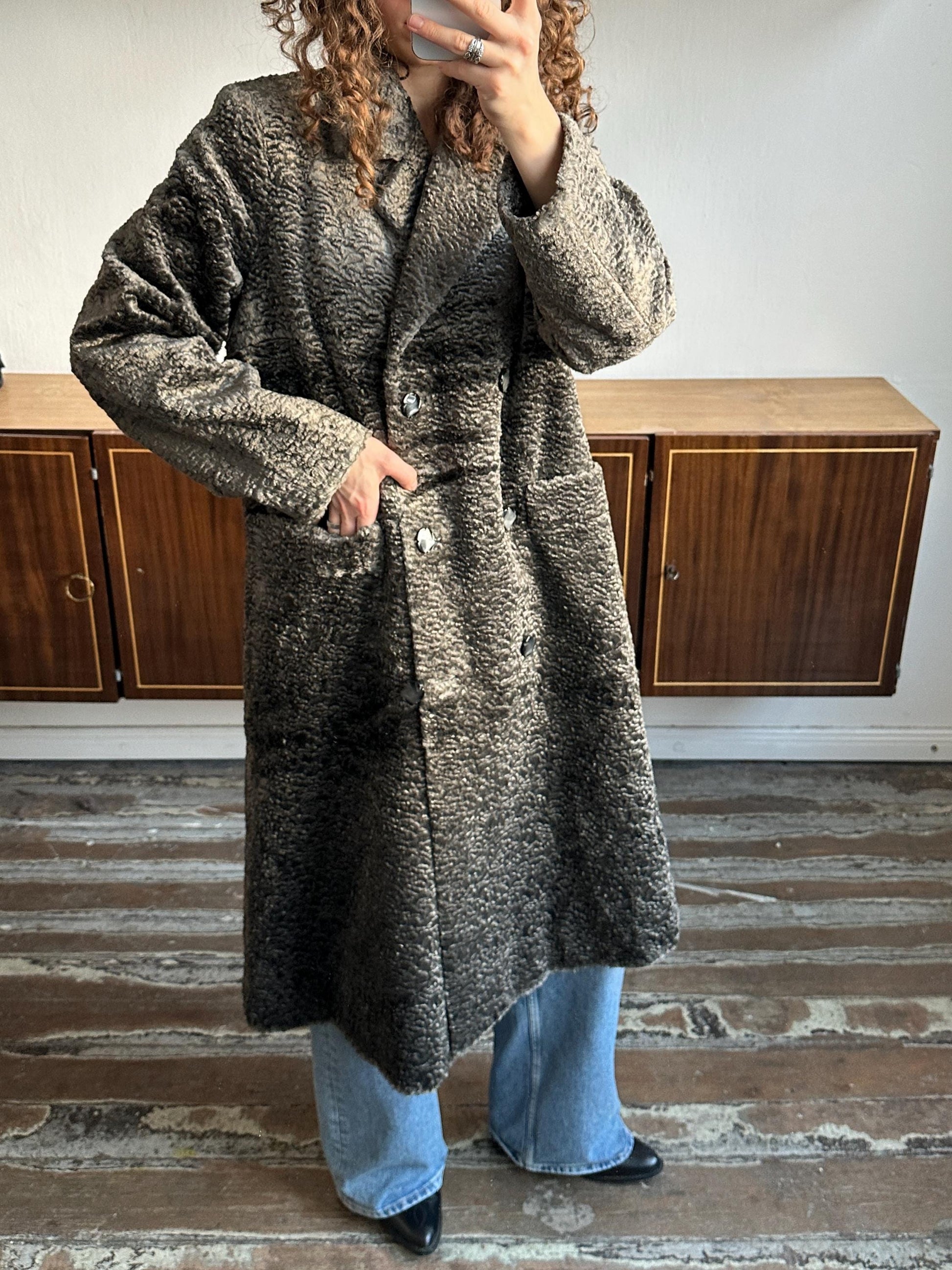 A person wearing a long gray coat is taking a selfie in front of a wooden cabinet.