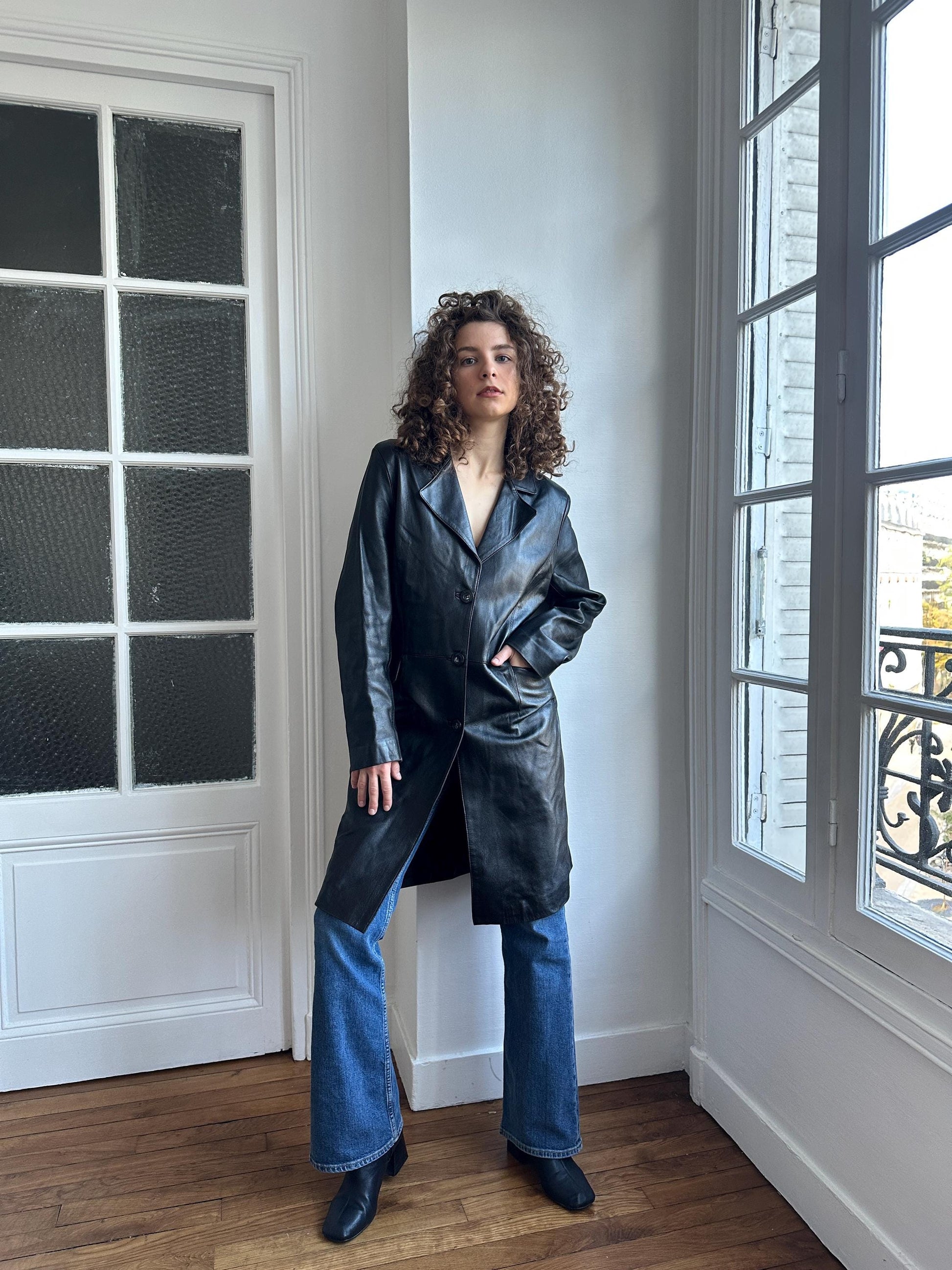 A woman in a black leather coat stands in a room with a large window, looking directly at the camera.