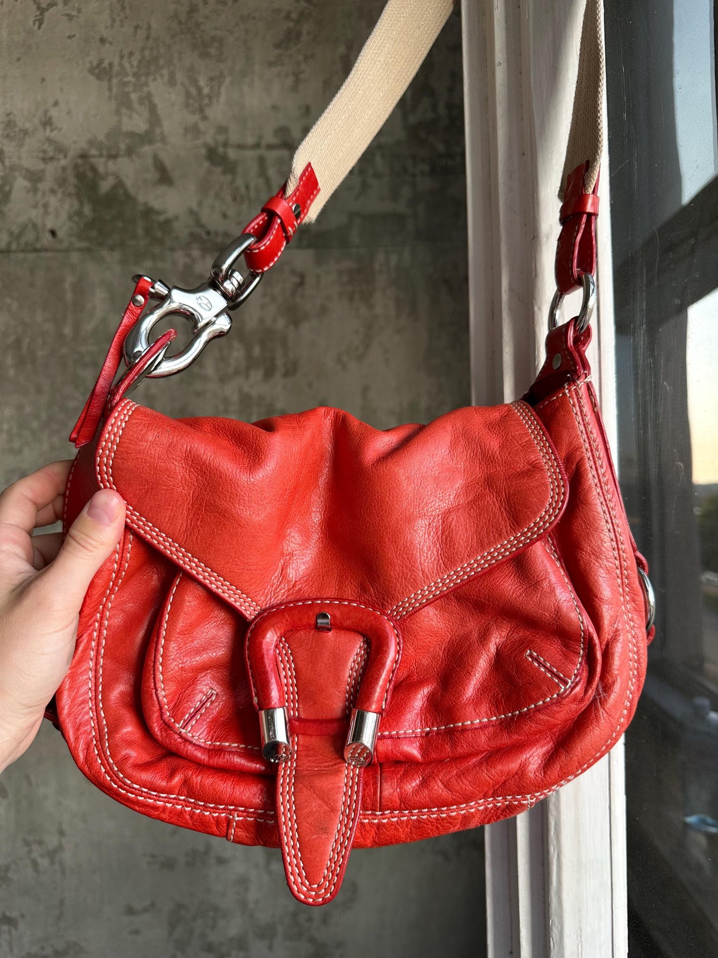 A hand is holding a red leather handbag with a metal clasp and a beige strap.