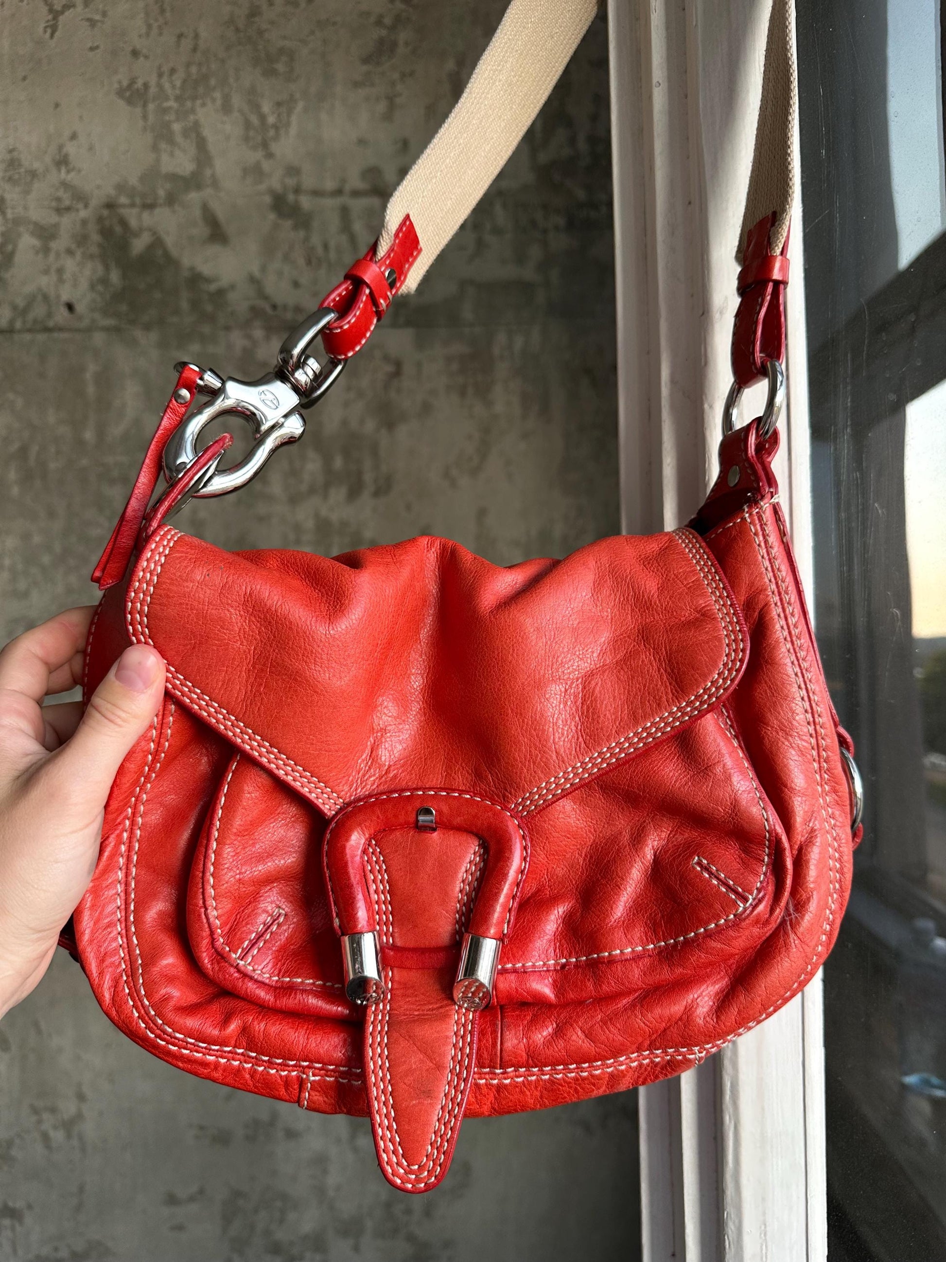 A hand is holding a red leather handbag with a metal clasp and a beige strap.