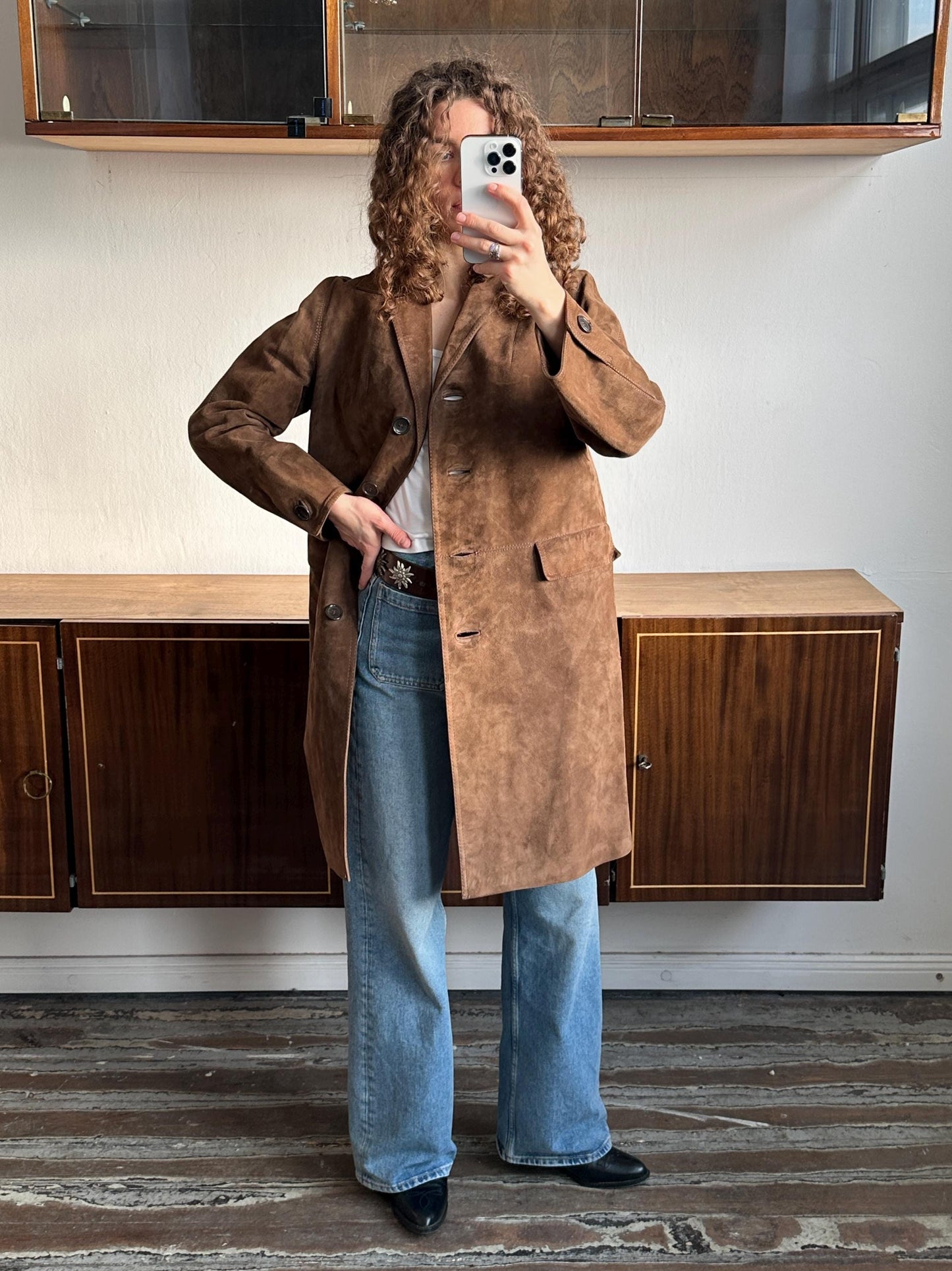 A person is taking a selfie with a brown coat and blue jeans, standing in front of a wooden cabinet.