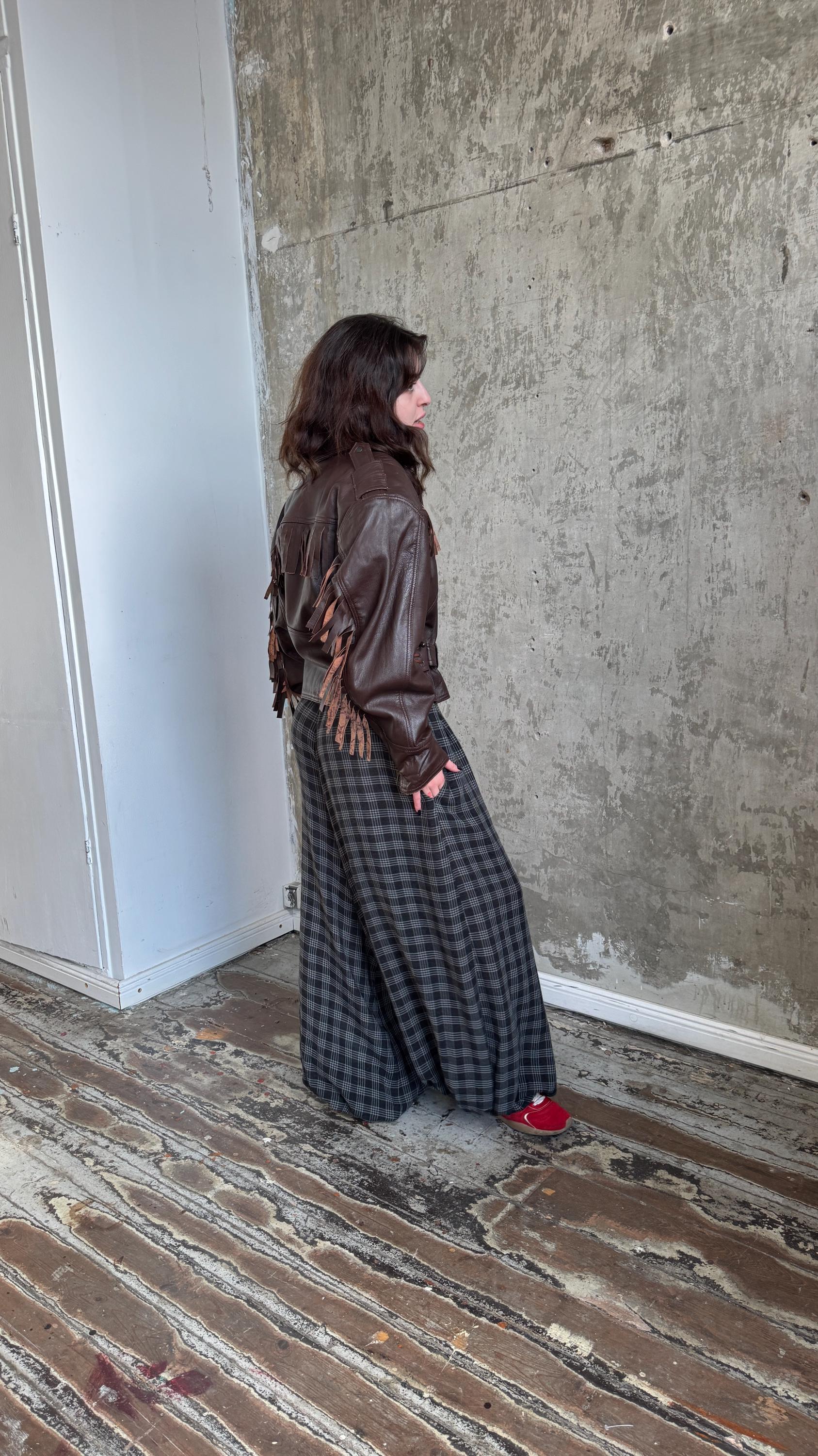 A person wearing a brown leather jacket and a long plaid skirt stands in a room with a concrete wall.