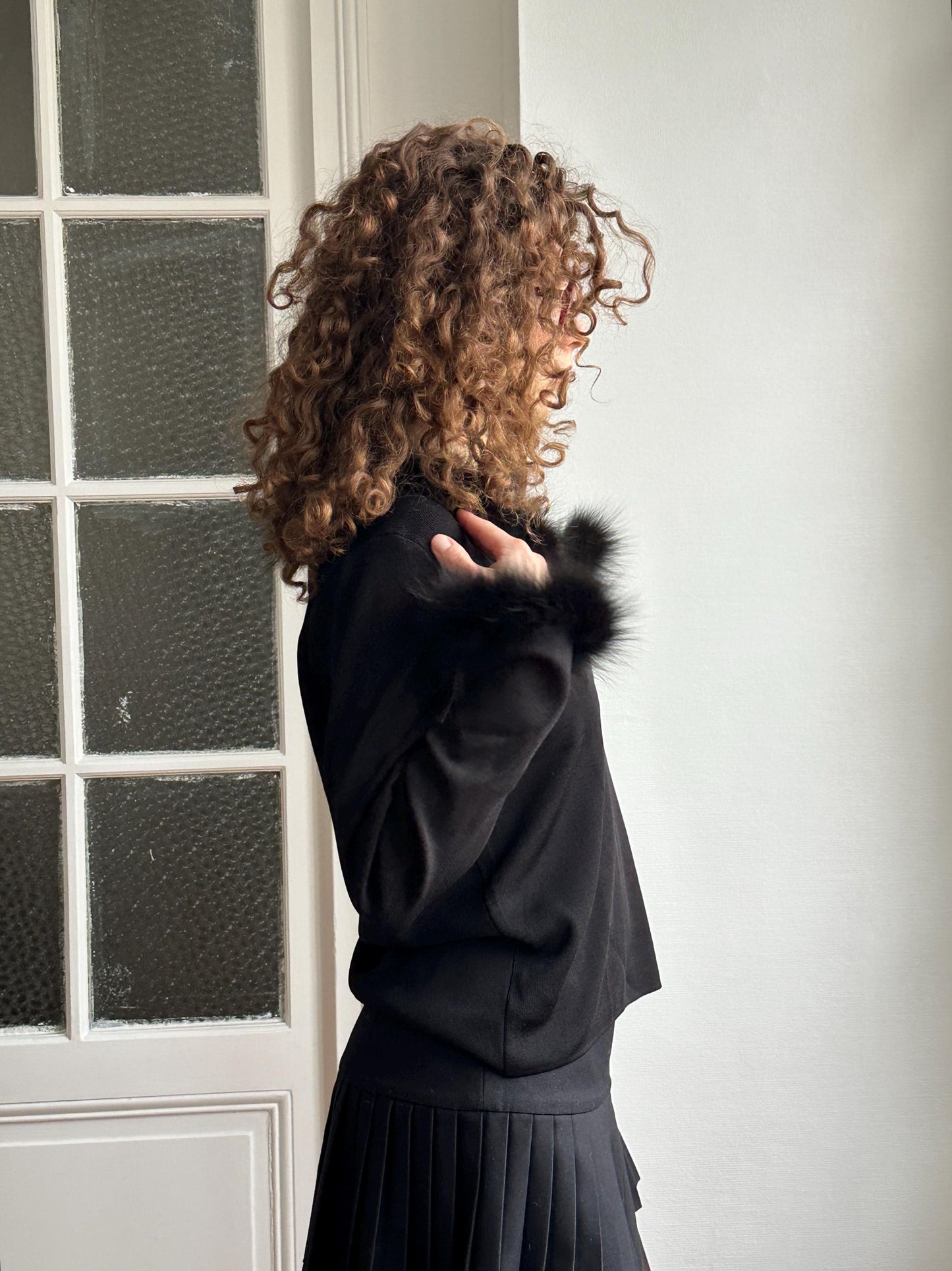 A woman with curly hair stands in front of a window, wearing a black outfit with fur trim.