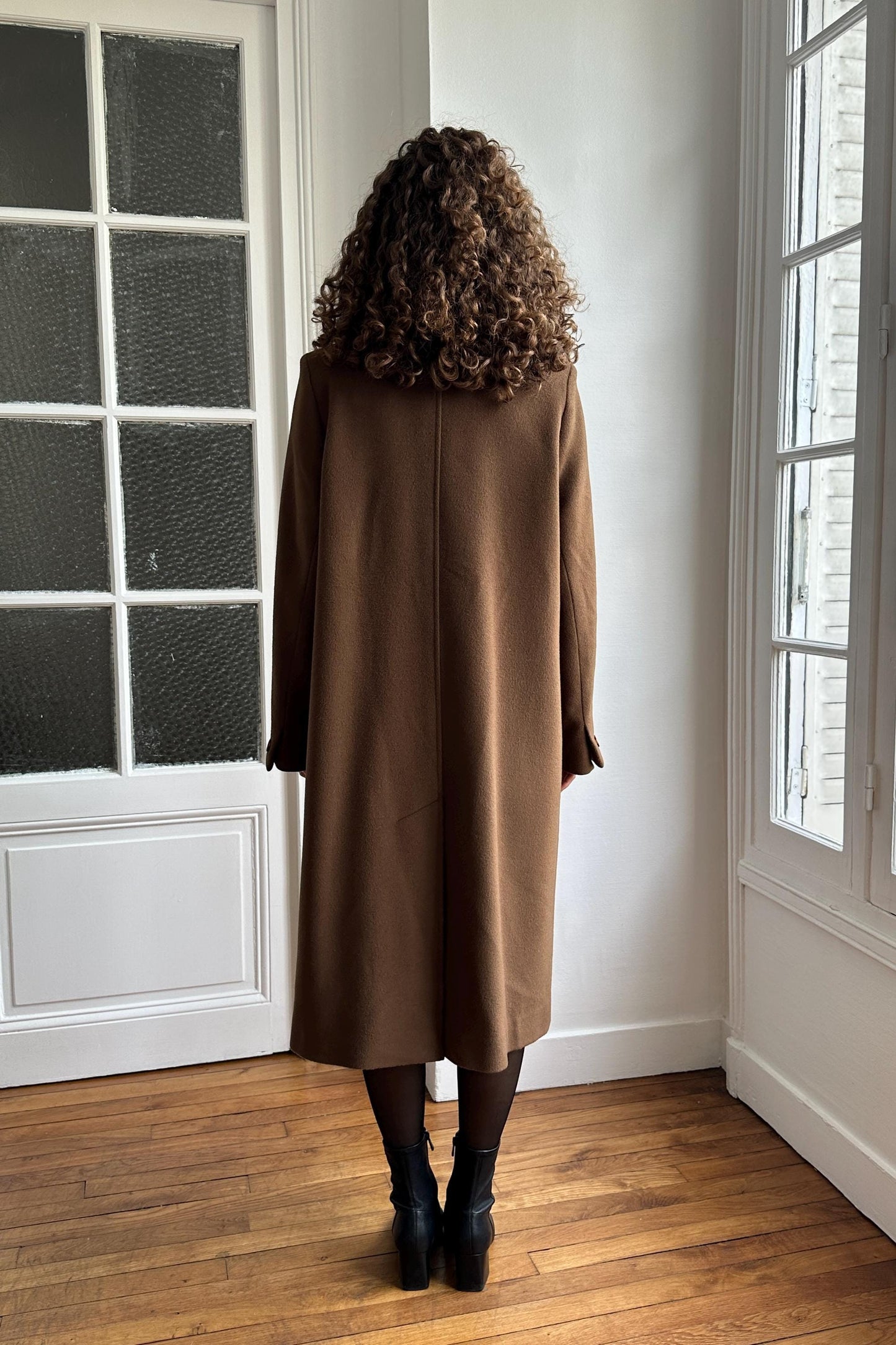 A person with curly hair wearing a brown coat stands in a room with white walls and a window.