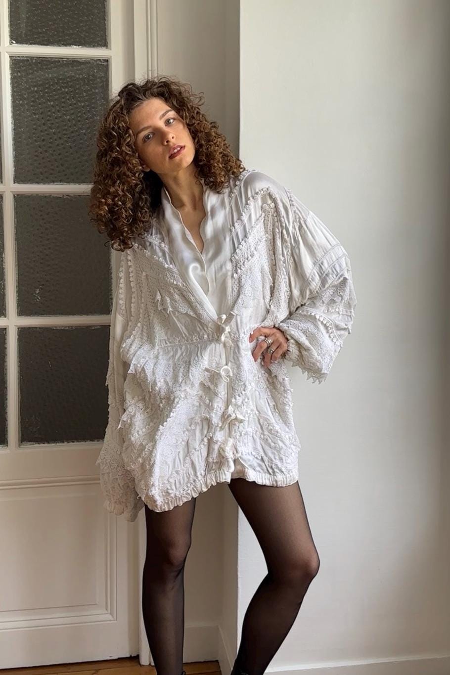 A woman with curly hair wearing a white, ruffled dress stands in front of a white wall, posing with a thoughtful expression.
