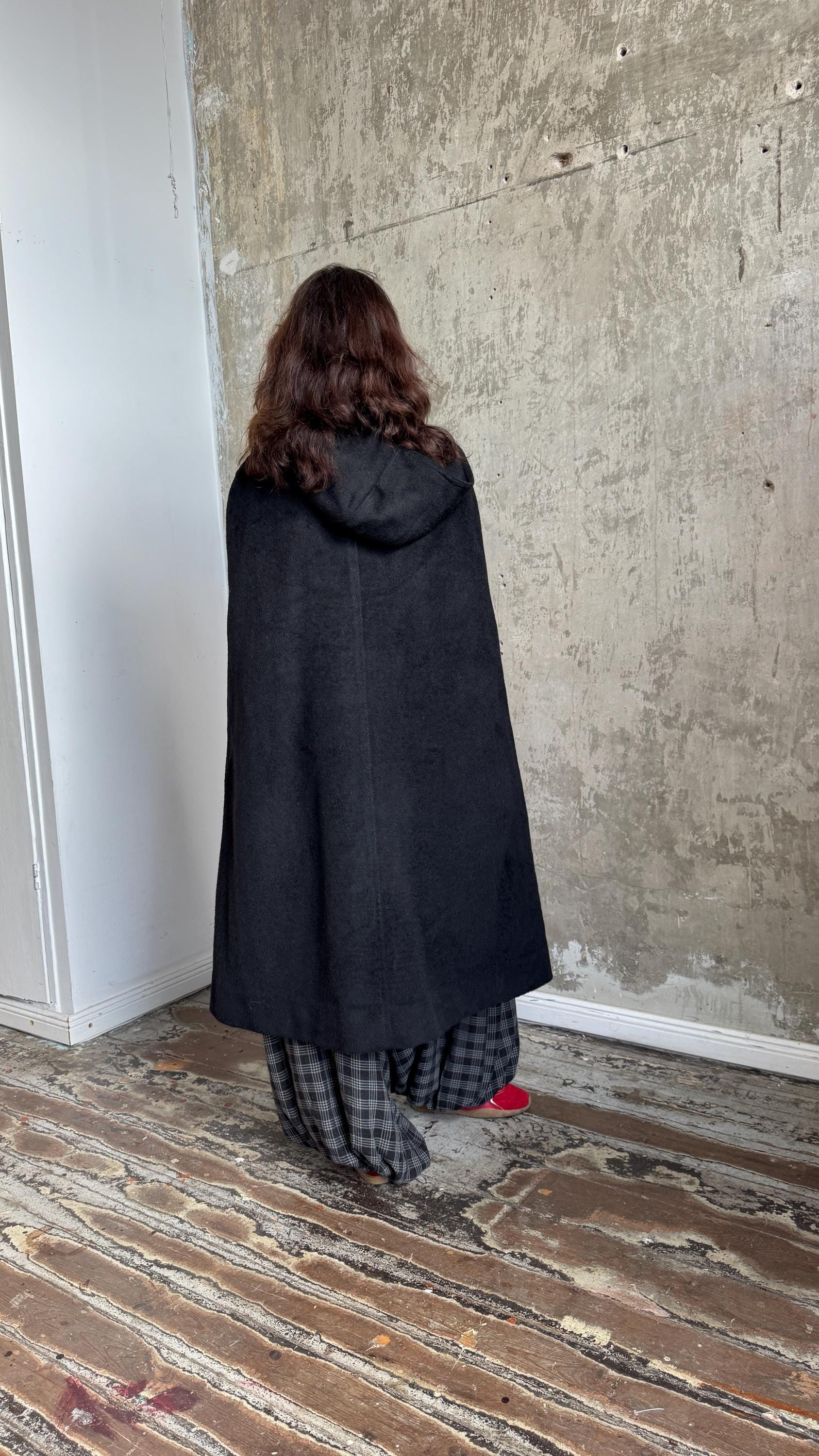 A person wearing a black cape is kneeling on a wooden floor in front of a concrete wall.