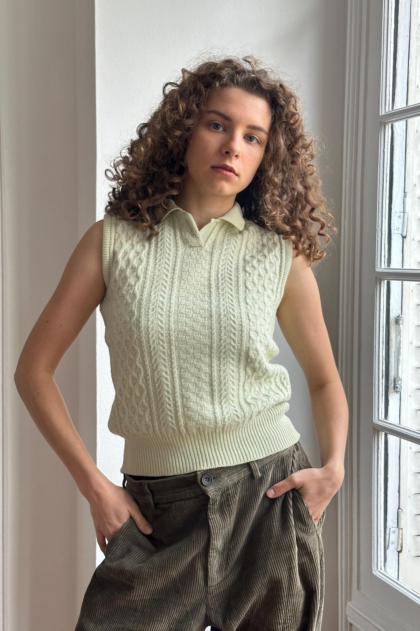 A young woman with curly hair stands in front of a window, wearing a white sleeveless sweater and brown pants.