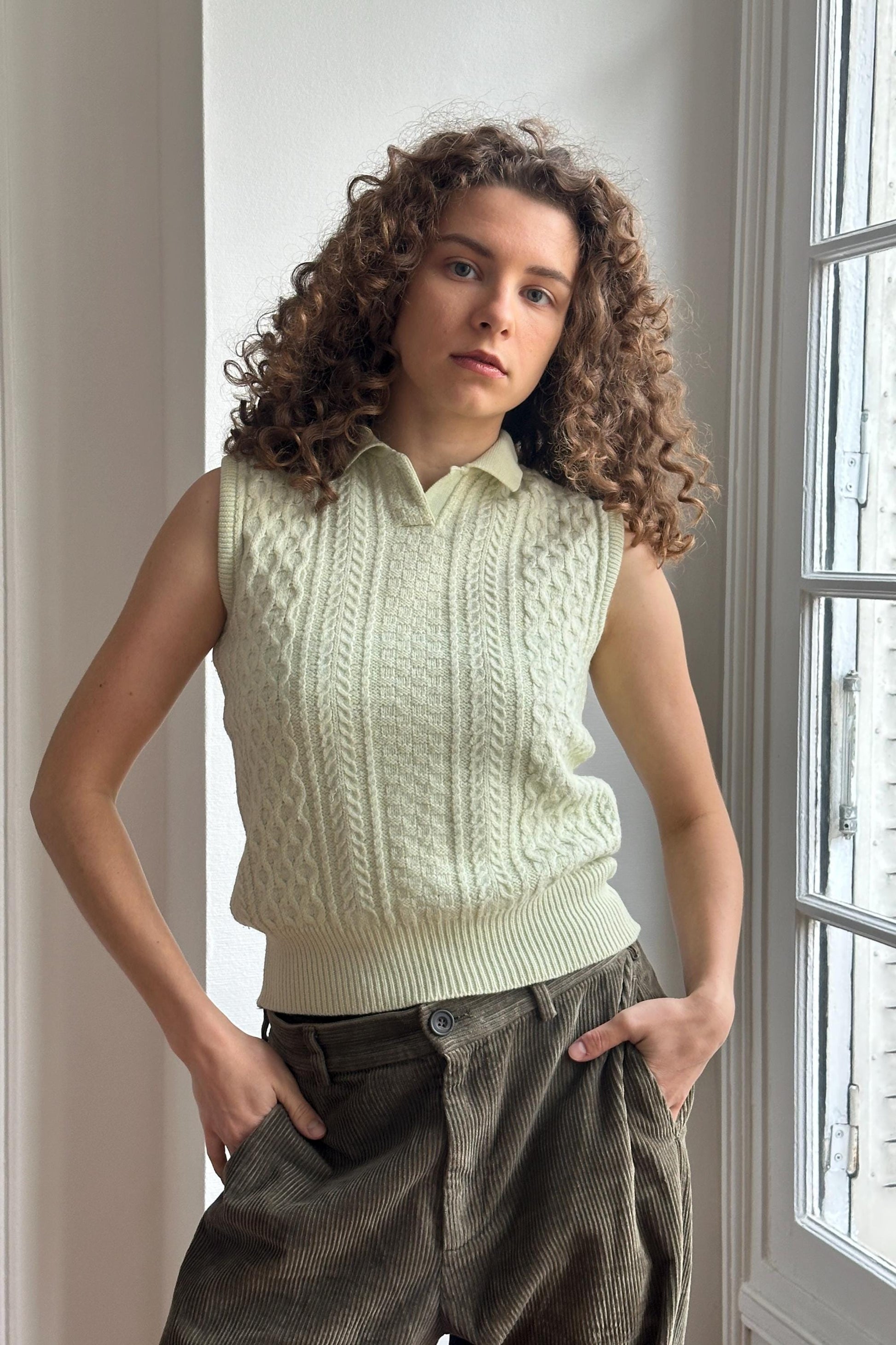 A young woman with curly hair stands in front of a window, wearing a white sleeveless sweater and brown pants.
