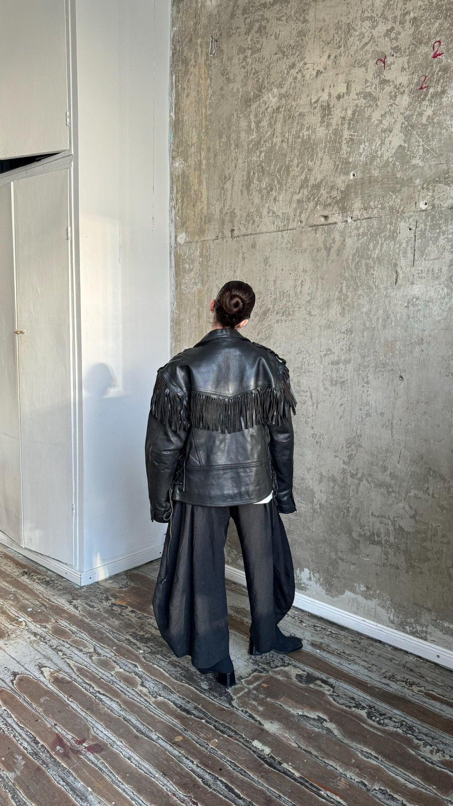 A person wearing a black leather jacket with fringe stands in a room with a concrete wall and wooden floor.