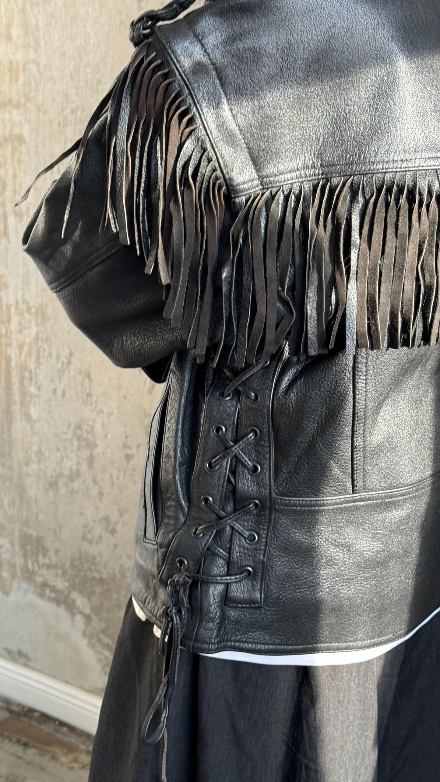 A close-up view of a black leather jacket with fringe detailing, including a cinched waist and a lace-up front.