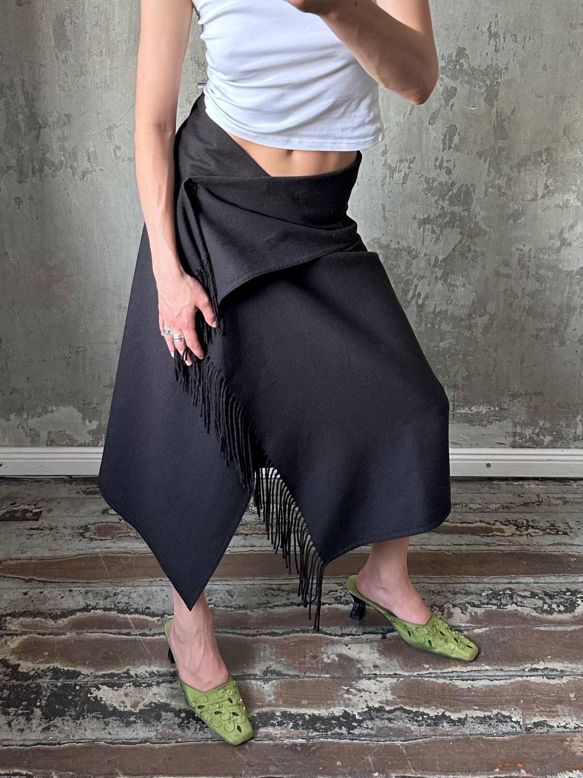 A person wearing a black skirt with fringe details and green high heels is standing in front of a concrete wall.