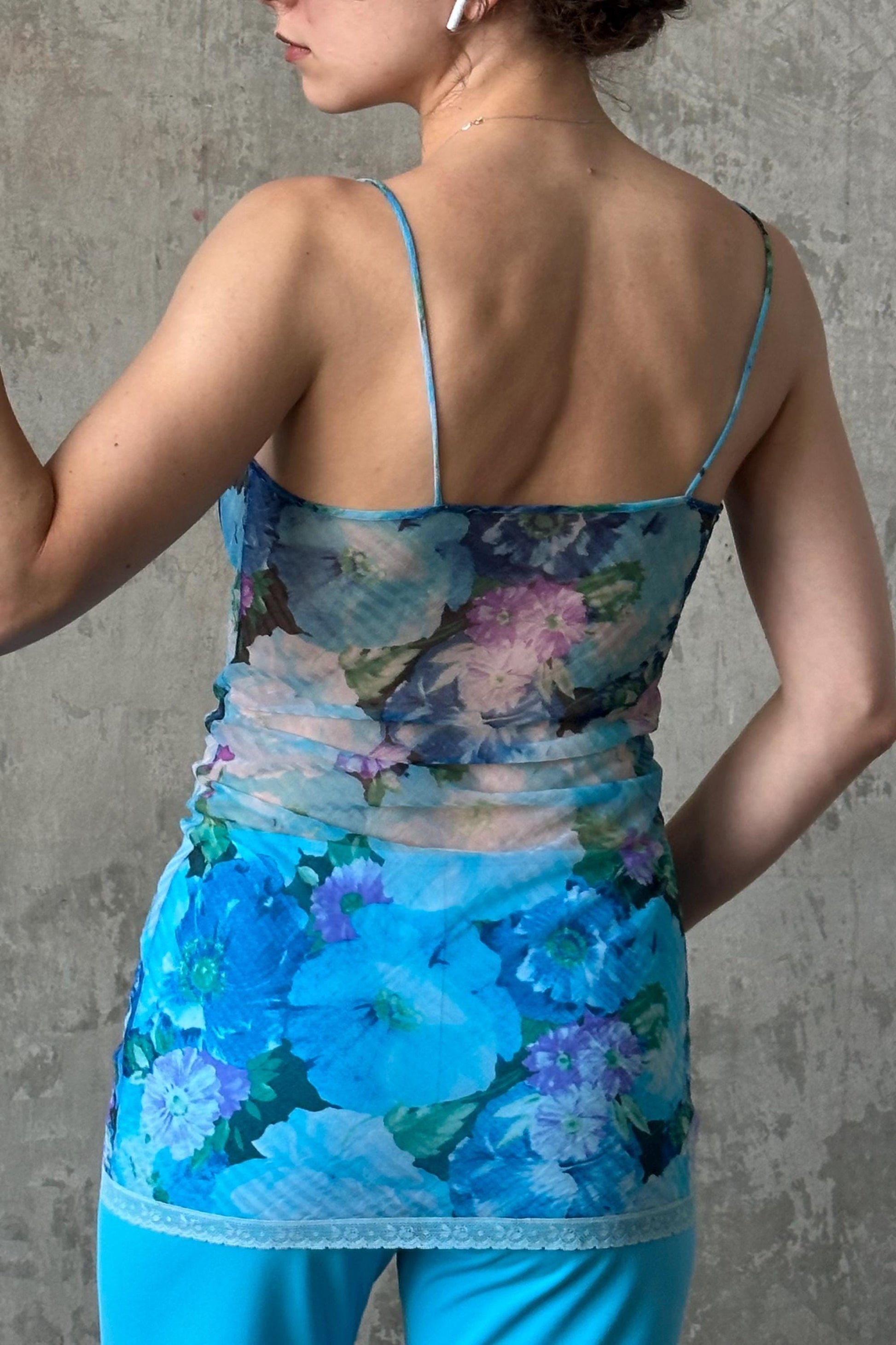 A woman wearing a blue floral print top and matching blue pants stands against a concrete wall.