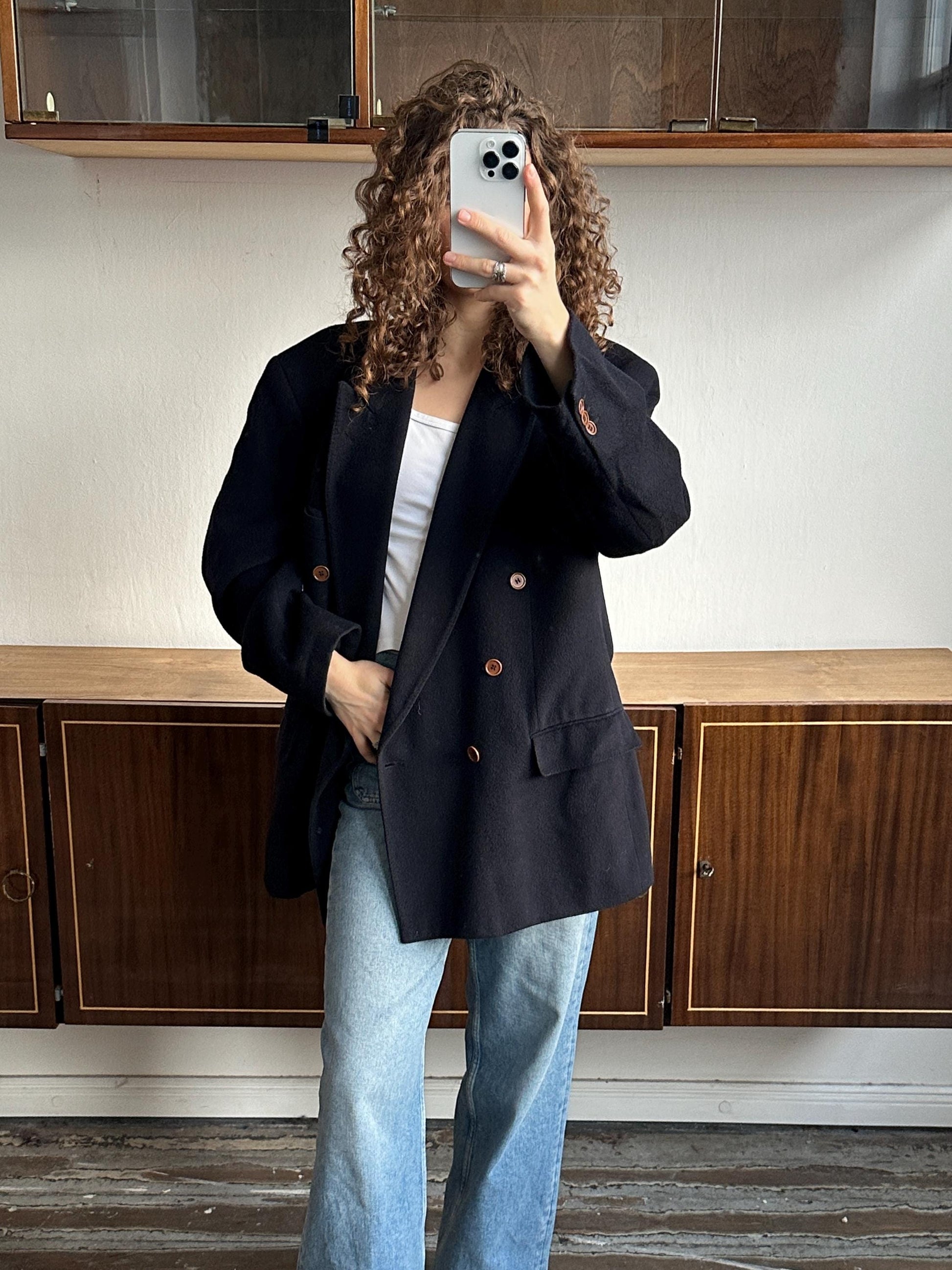 A person with curly hair is taking a selfie with a smartphone, wearing a black blazer and blue jeans.