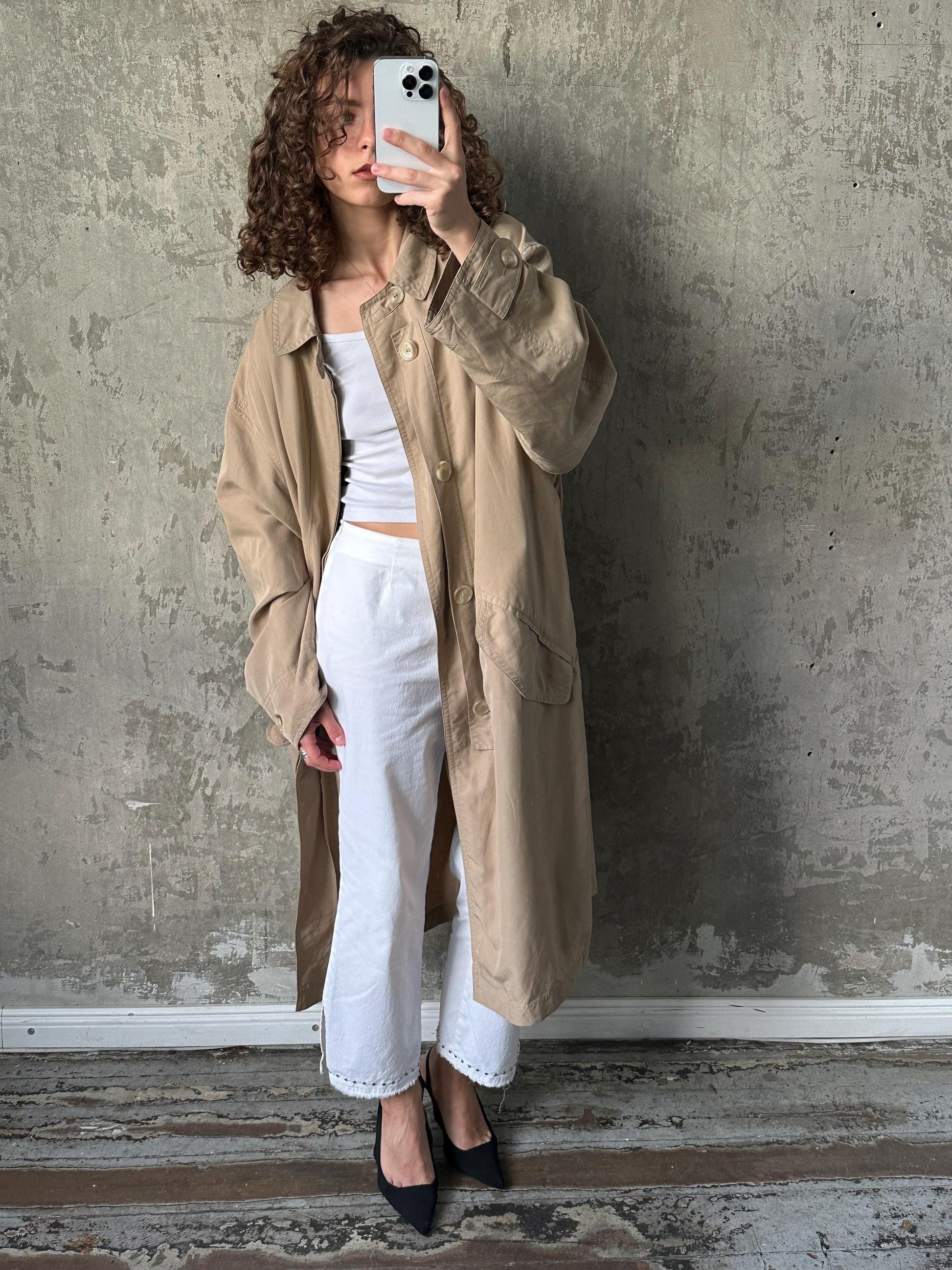A woman with curly hair is taking a selfie with her phone, wearing a beige coat and white pants against a concrete wall.