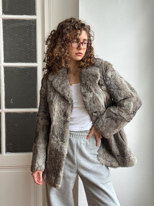 A woman with curly hair and glasses is standing in front of a white door, wearing a gray fur coat and gray sweatpants.
