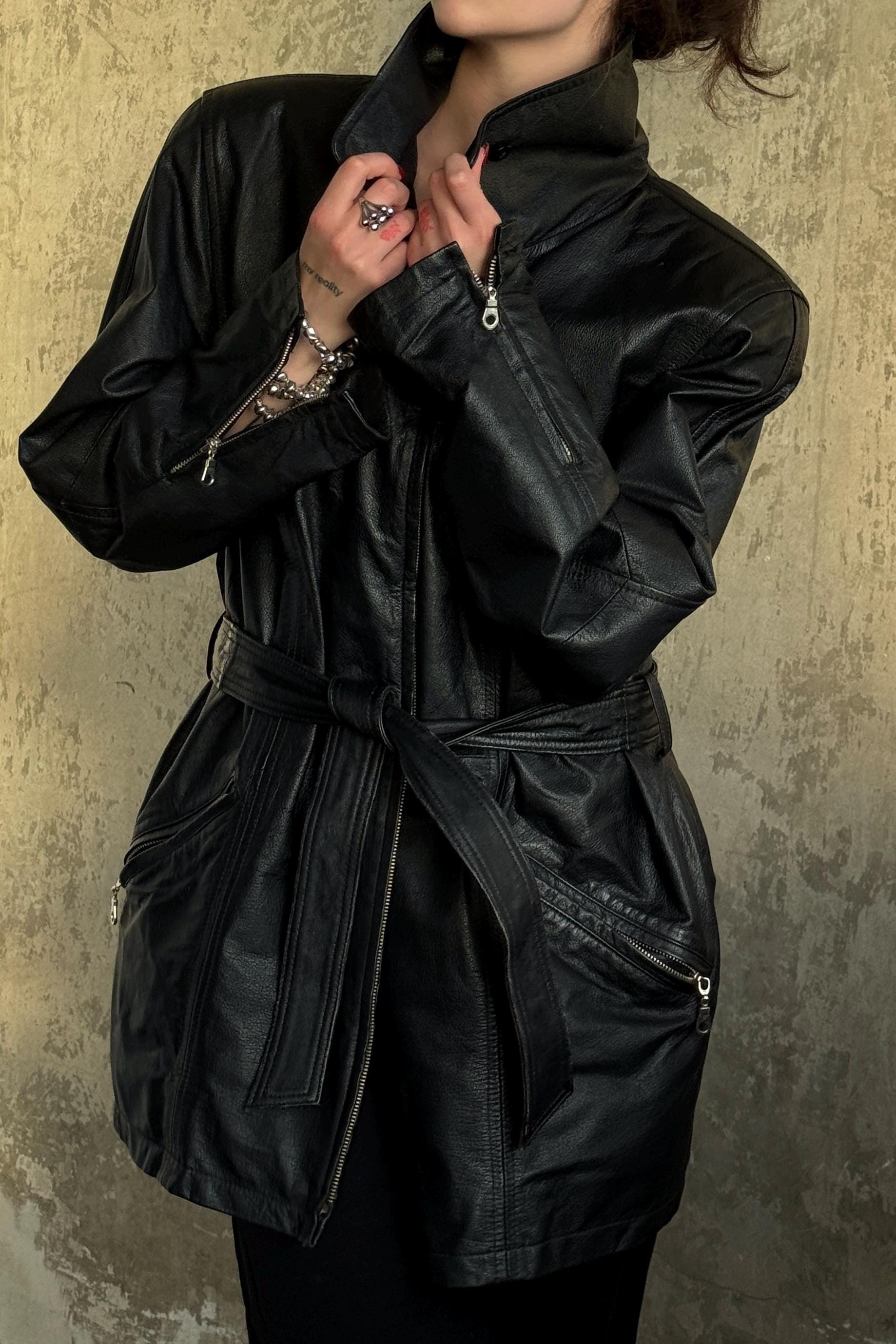 A person wearing a black leather jacket with a belt and zippers, standing against a textured wall.