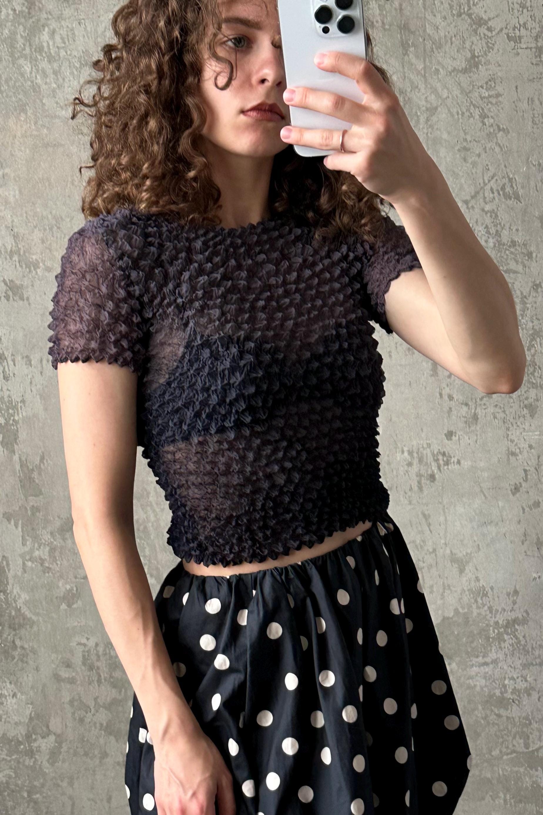 A woman with curly hair is taking a selfie with a white iPhone, wearing a black and white polka dot skirt and a black top with a ruffled texture.