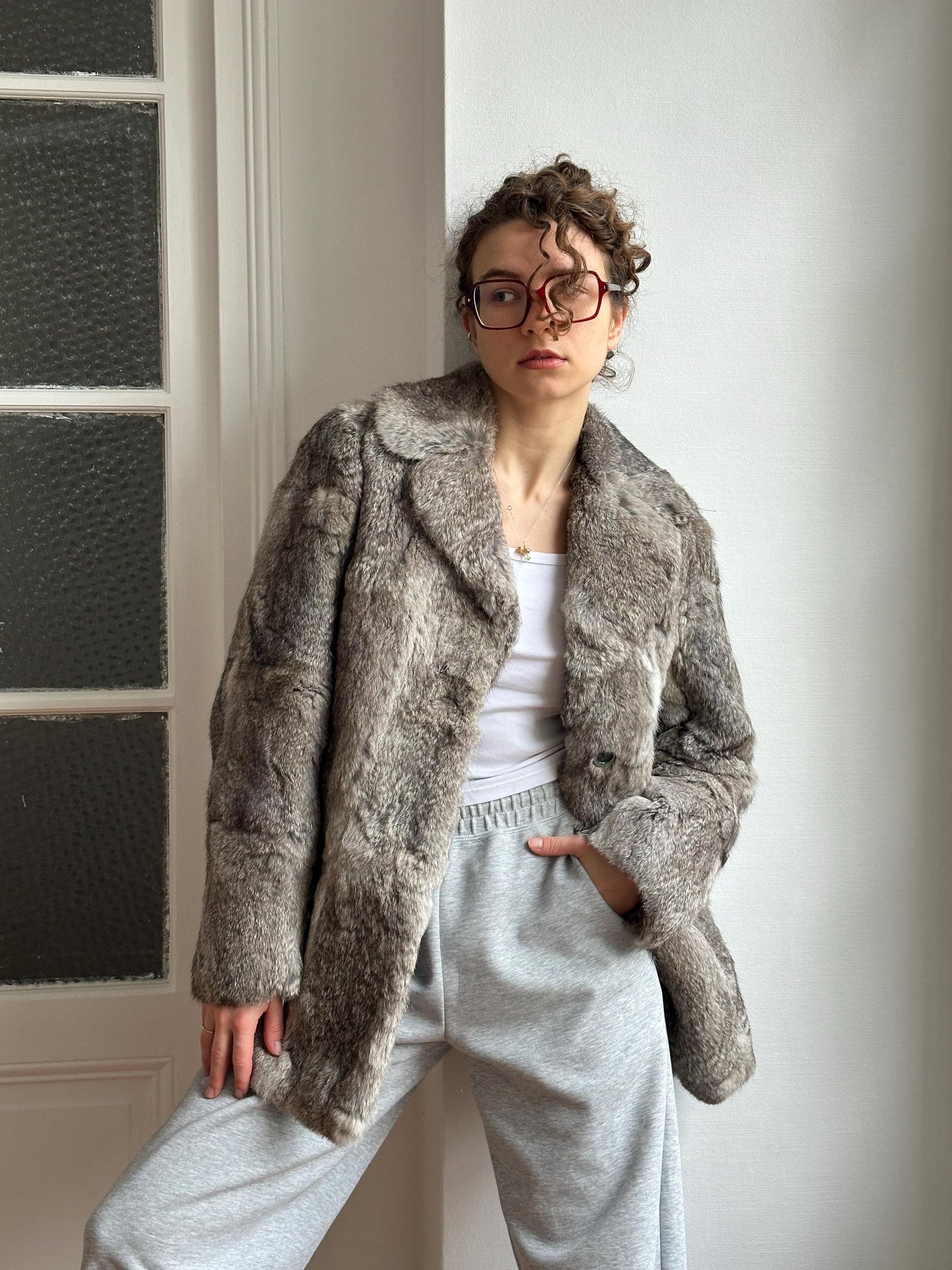 A person wearing a gray fur coat and glasses is leaning against a white wall.