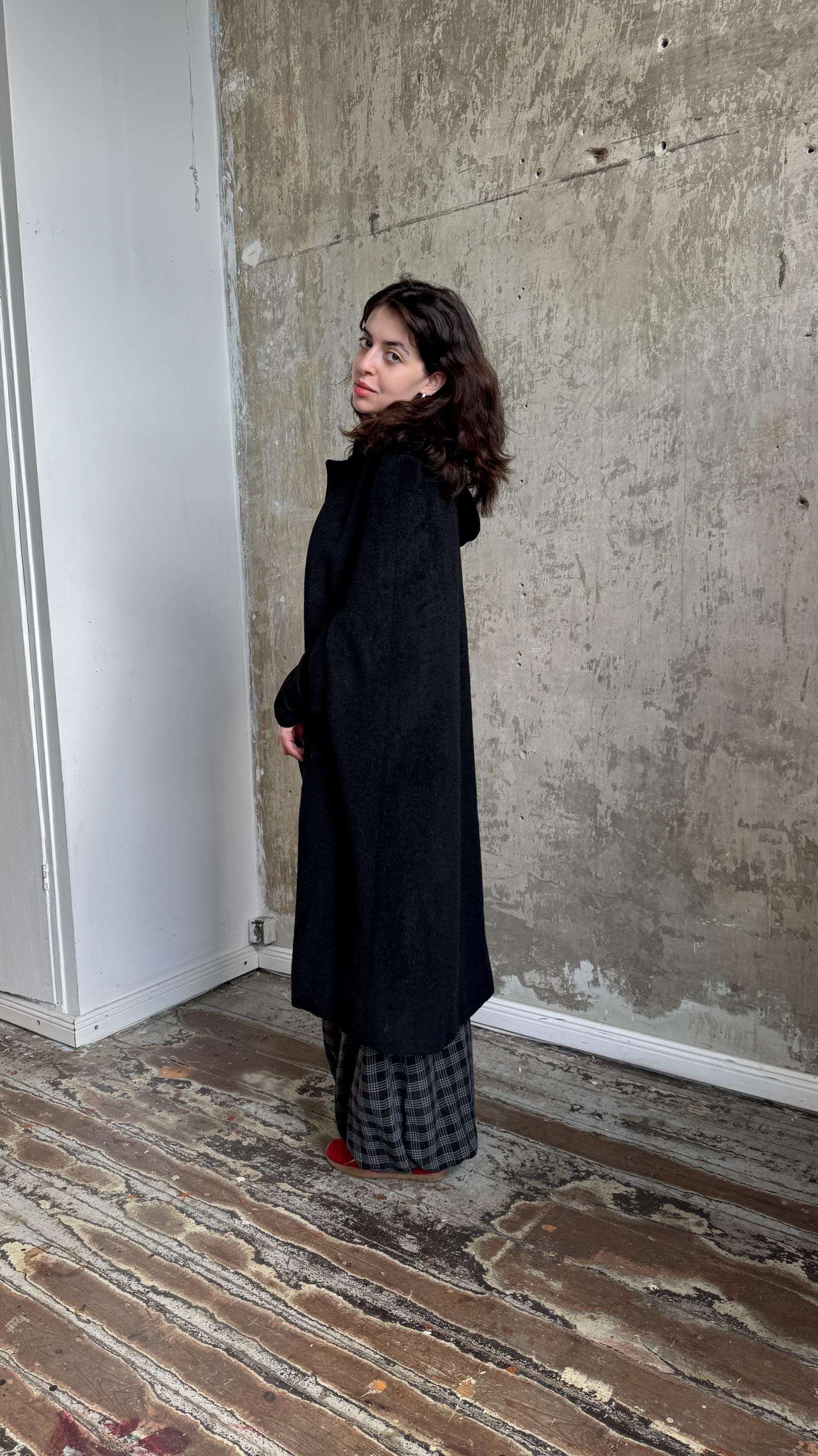 A woman in a black coat stands in a room with a concrete wall and wooden floor.