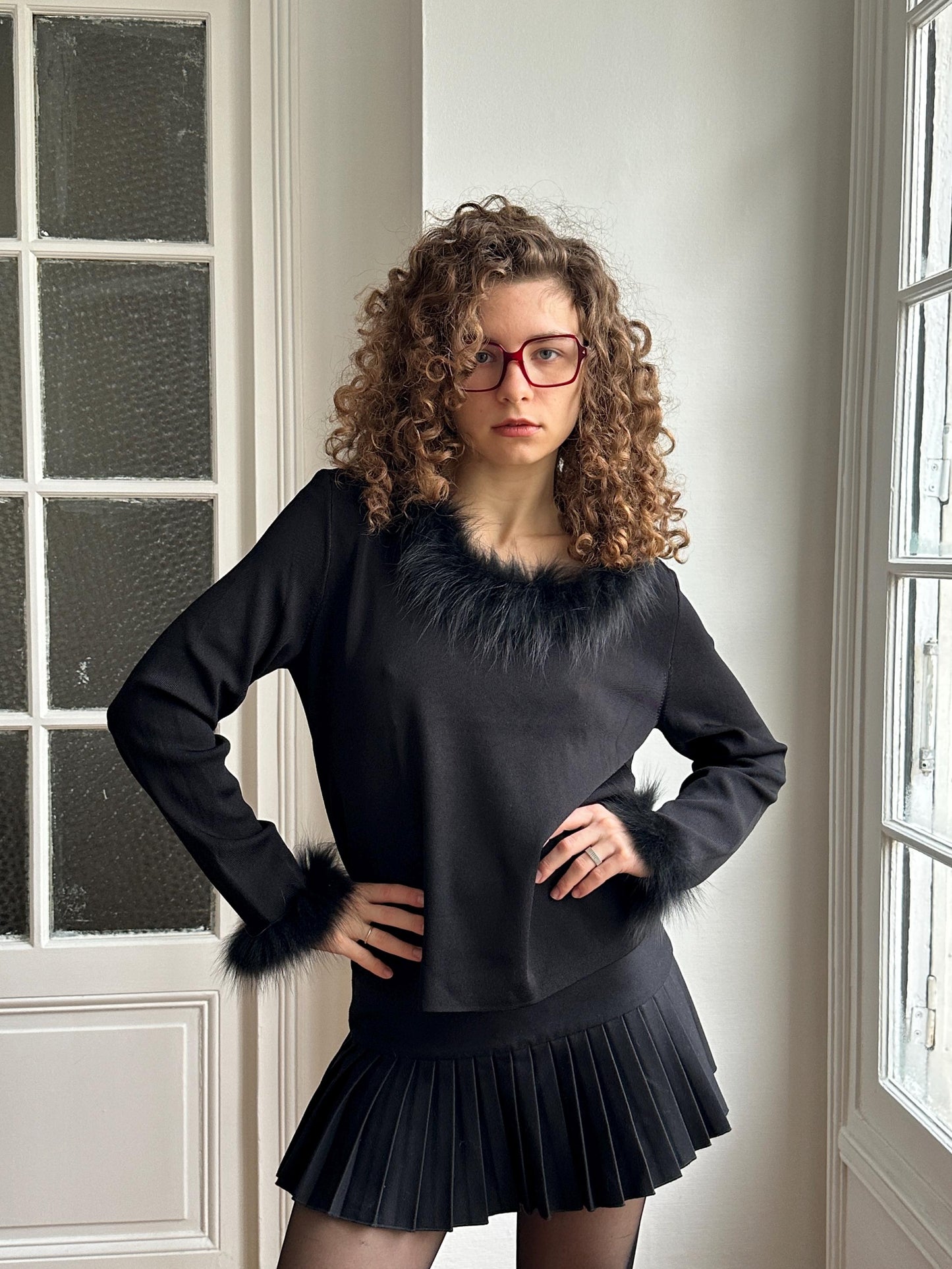 A woman with curly hair and glasses stands confidently in a black outfit with fur trim, posing in front of a window.