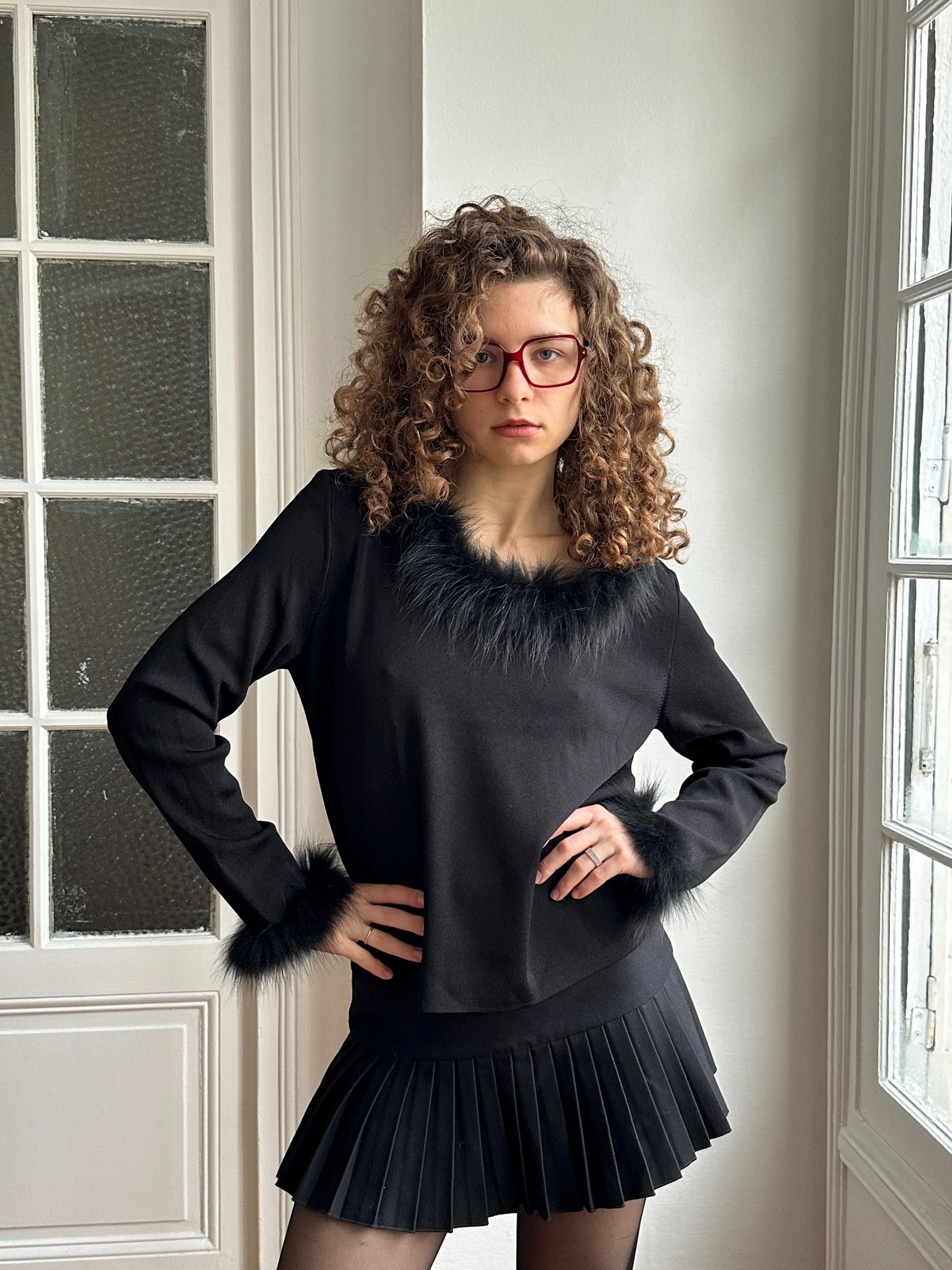 A woman with curly hair and glasses stands confidently in a black outfit with fur trim, posing in front of a window.