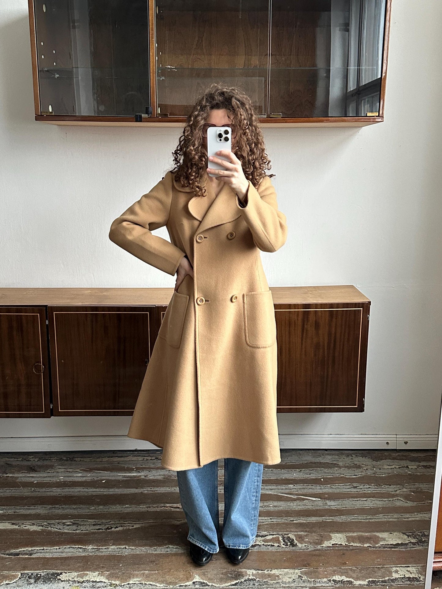 May include: A tan double-breasted coat with a notched collar, two rows of buttons, and two front pockets. The coat is knee-length and has an A-line shape. The person is wearing blue jeans.