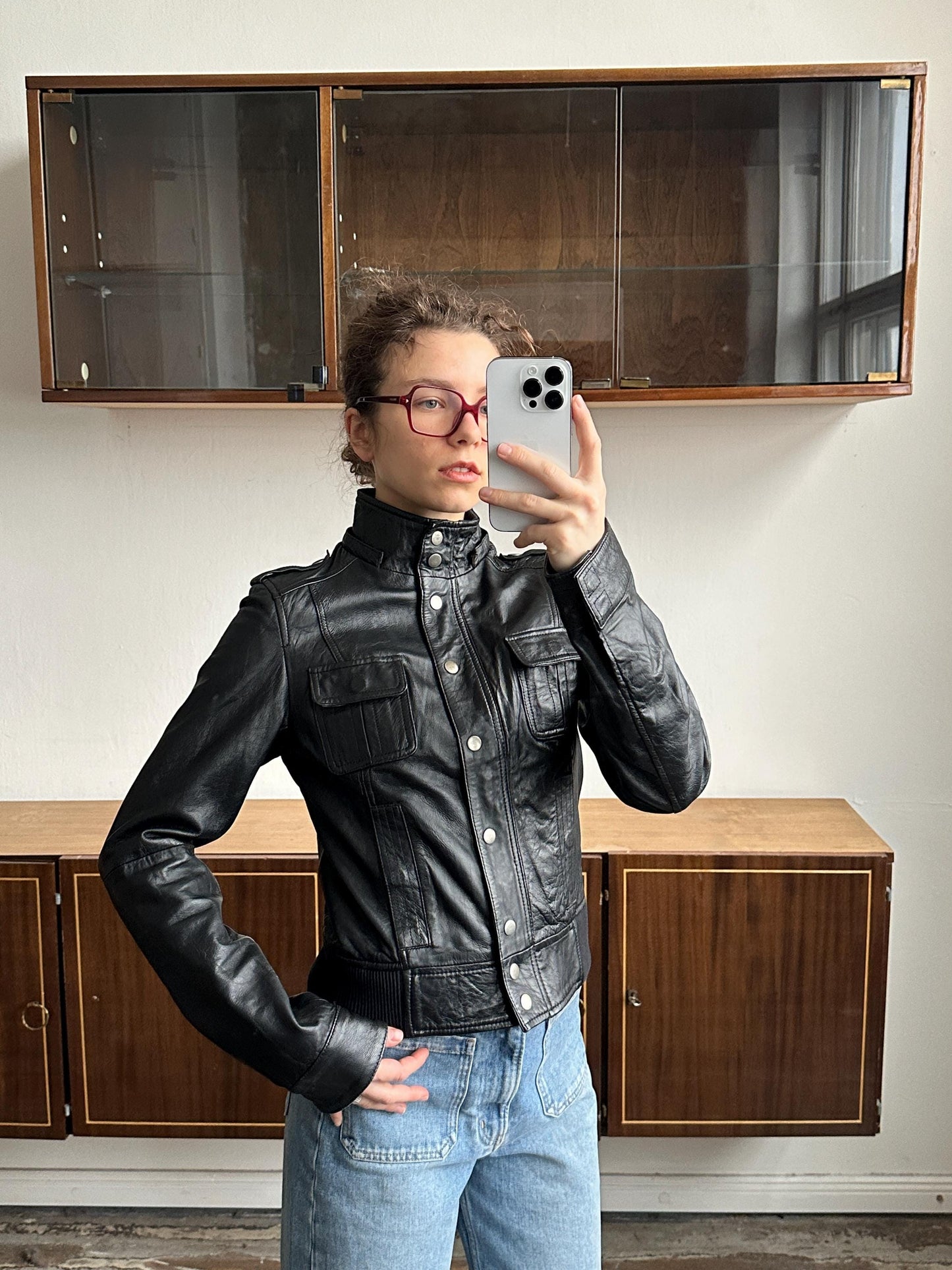 A person wearing a black leather jacket and red glasses is taking a selfie with a smartphone in front of a wooden cabinet.