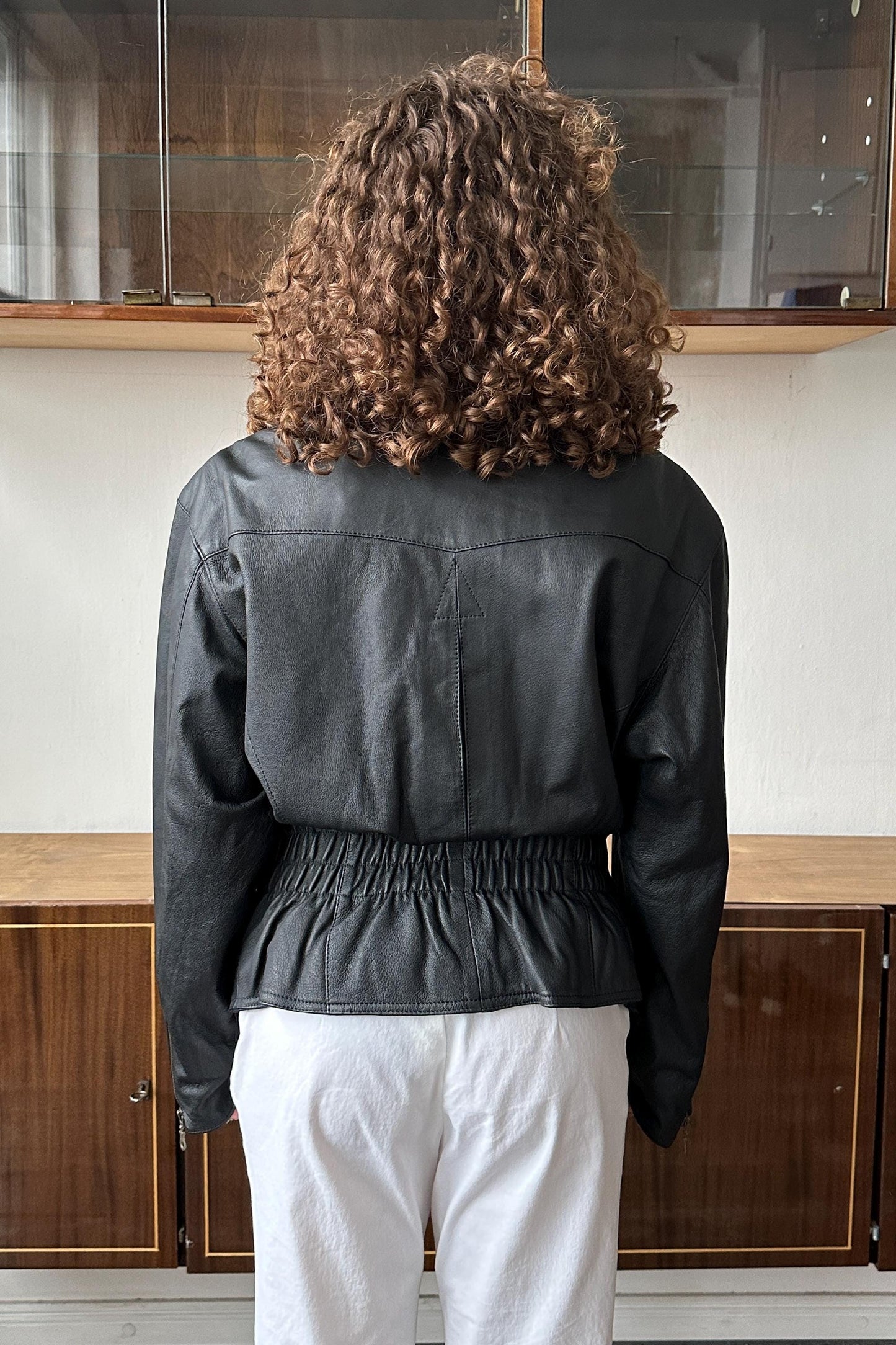 A person with curly hair wearing a black leather jacket and white pants stands facing away from the camera.