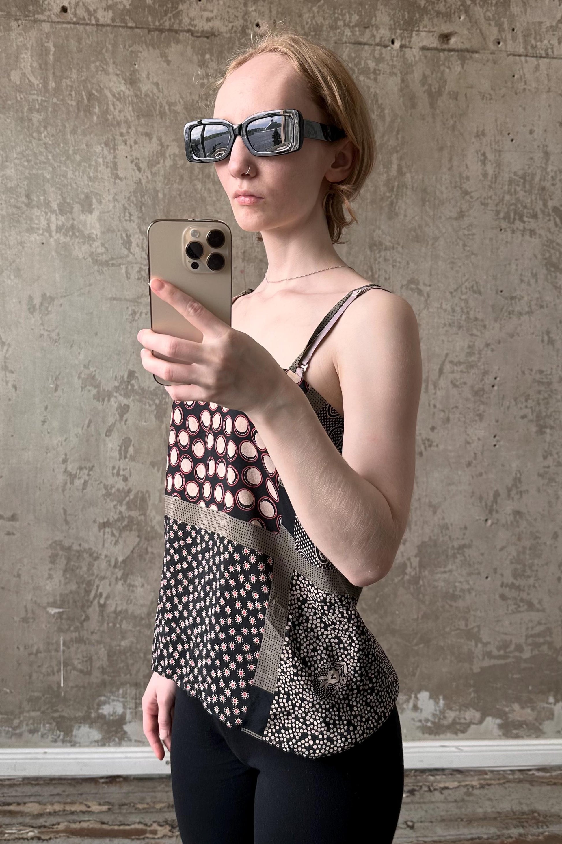 A woman wearing sunglasses and a patterned top is holding a smartphone and taking a selfie in front of a concrete wall.