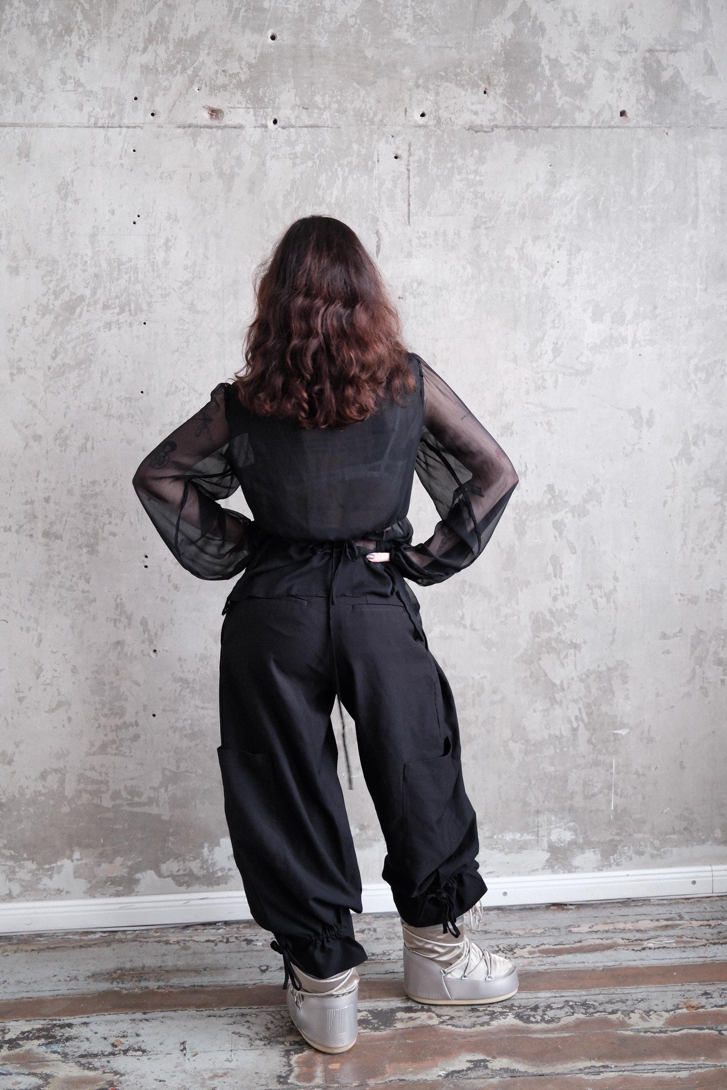 A person wearing black pants and a black top stands in front of a white wall, with their back turned to the camera.