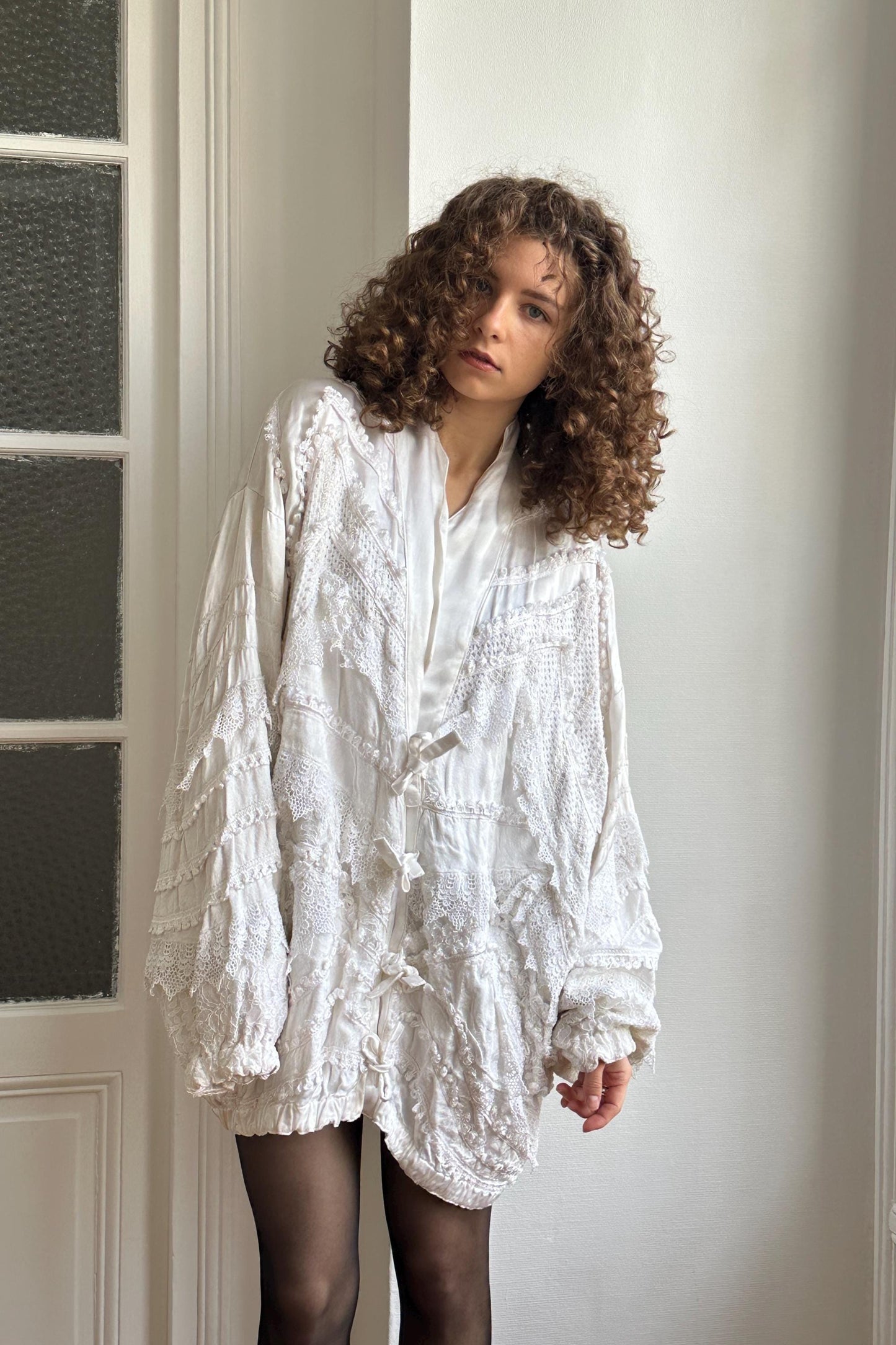 A young woman with curly hair wearing a white, ruffled jacket stands in a room with a white wall and a window.