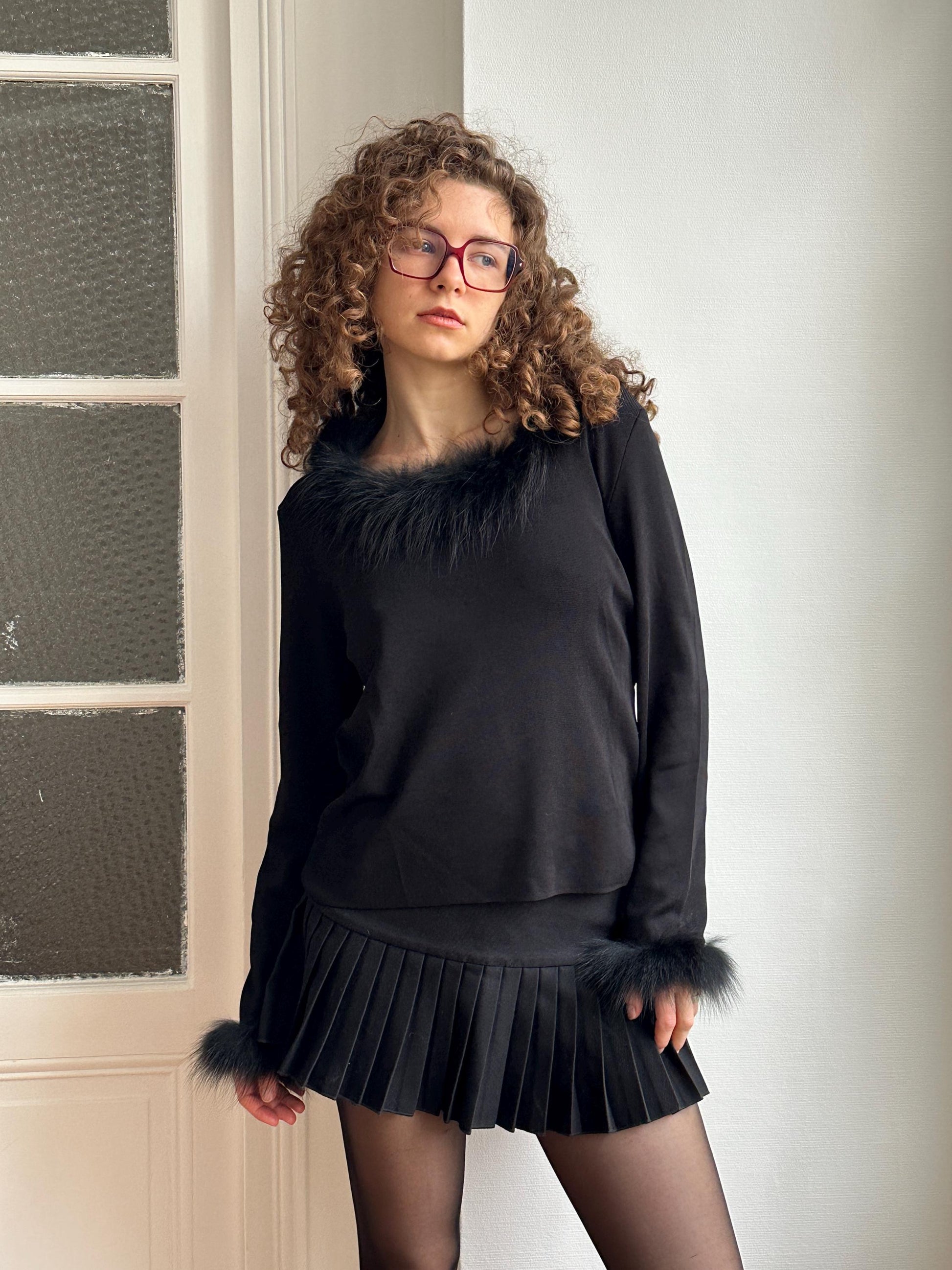 A young woman with curly hair and glasses is wearing a black outfit with a fur collar and skirt, posing in front of a white door.