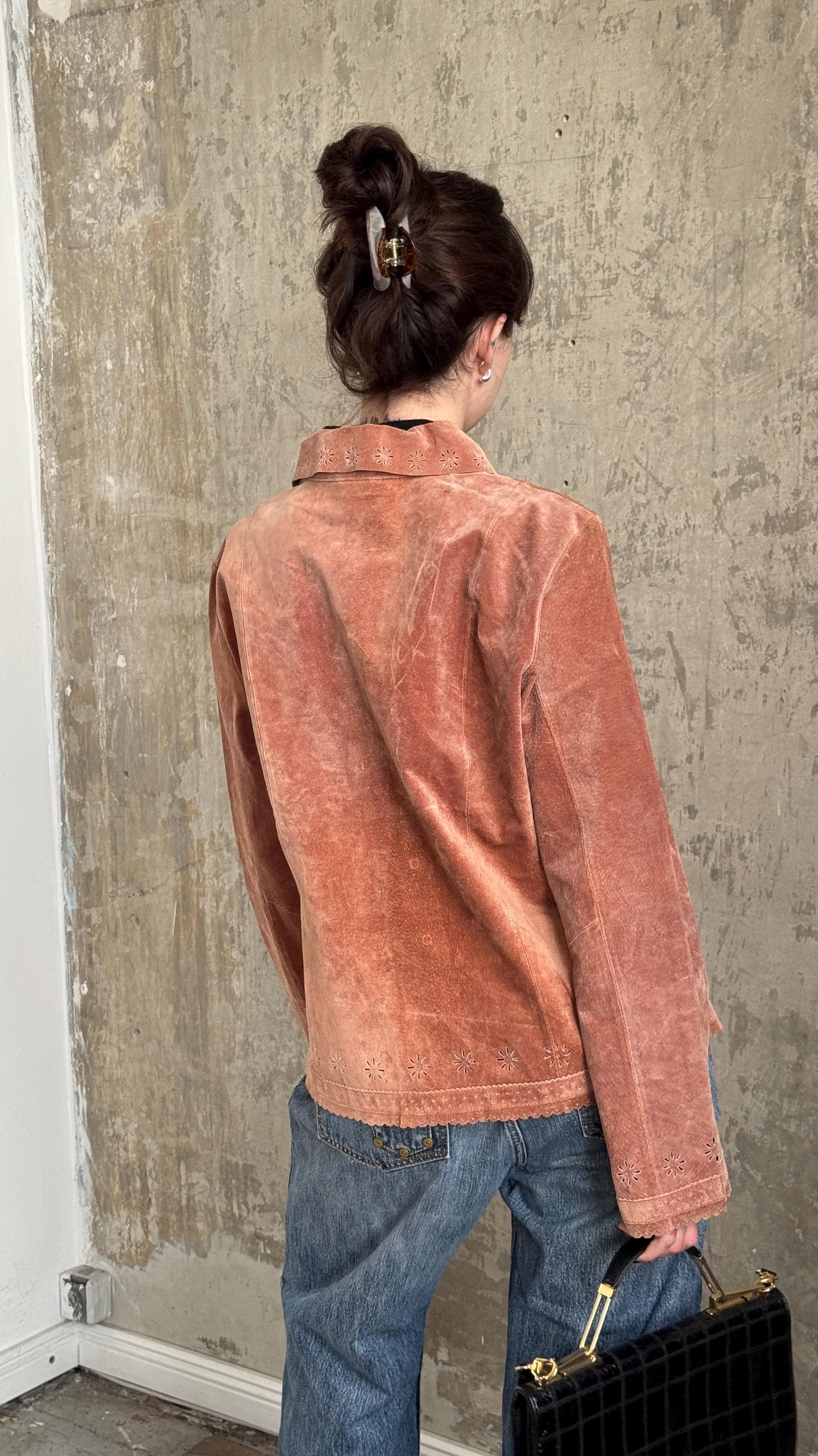 A woman wearing a pink jacket and jeans is standing in front of a concrete wall, holding a black purse.