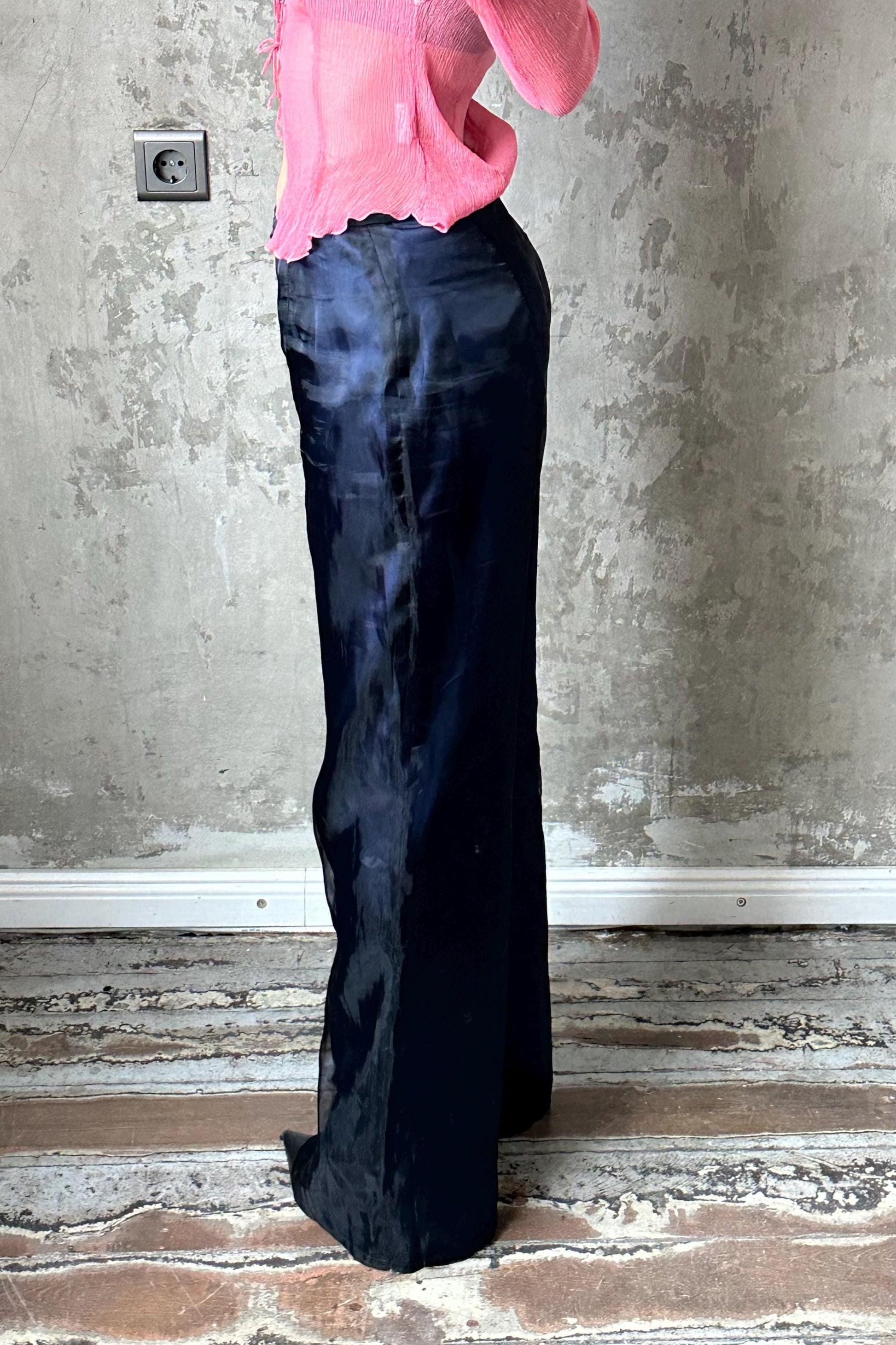 A person wearing a pink top and black pants stands in front of a concrete wall.