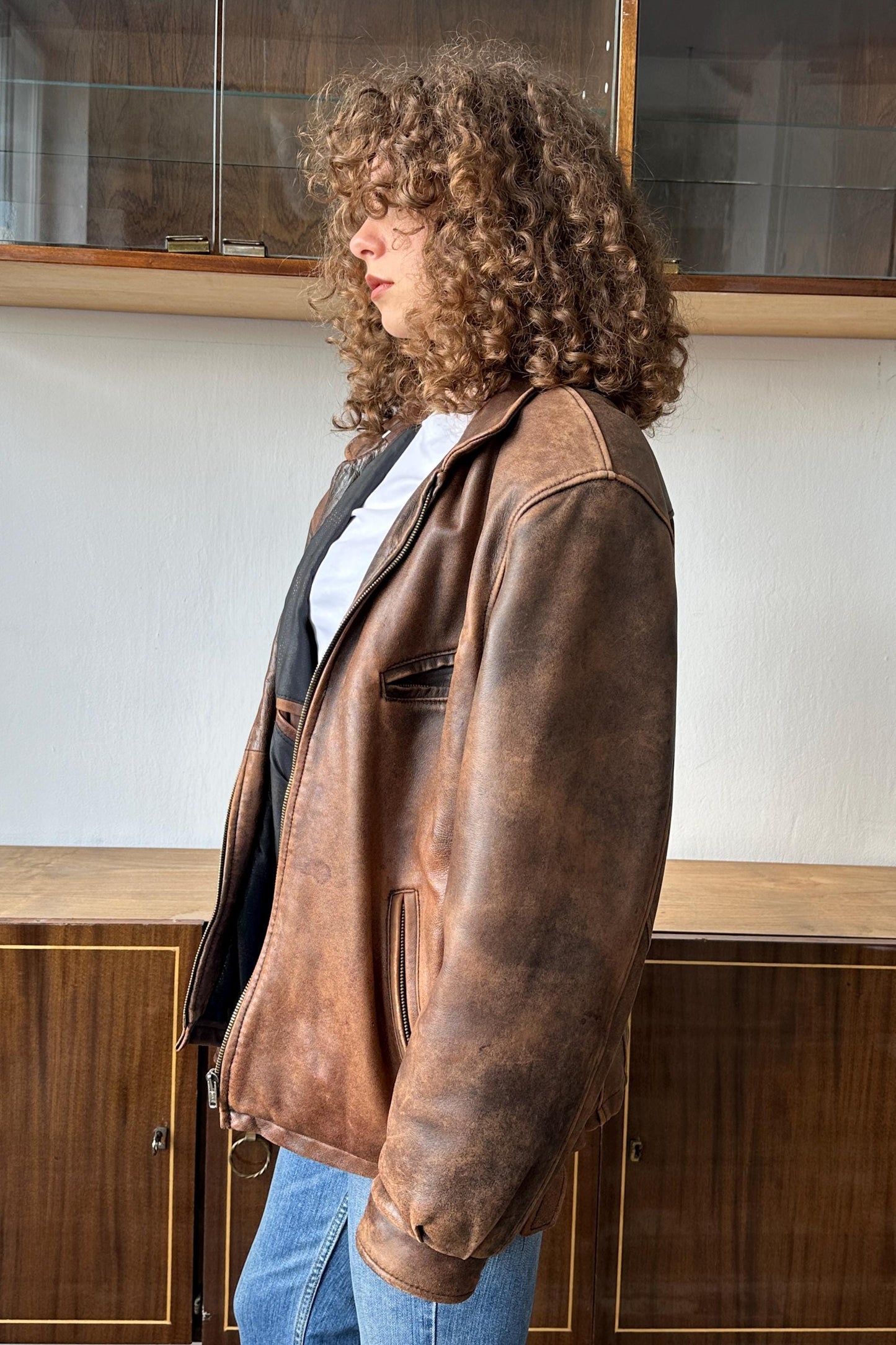A woman with curly hair wearing a brown leather jacket stands in front of a wooden cabinet.