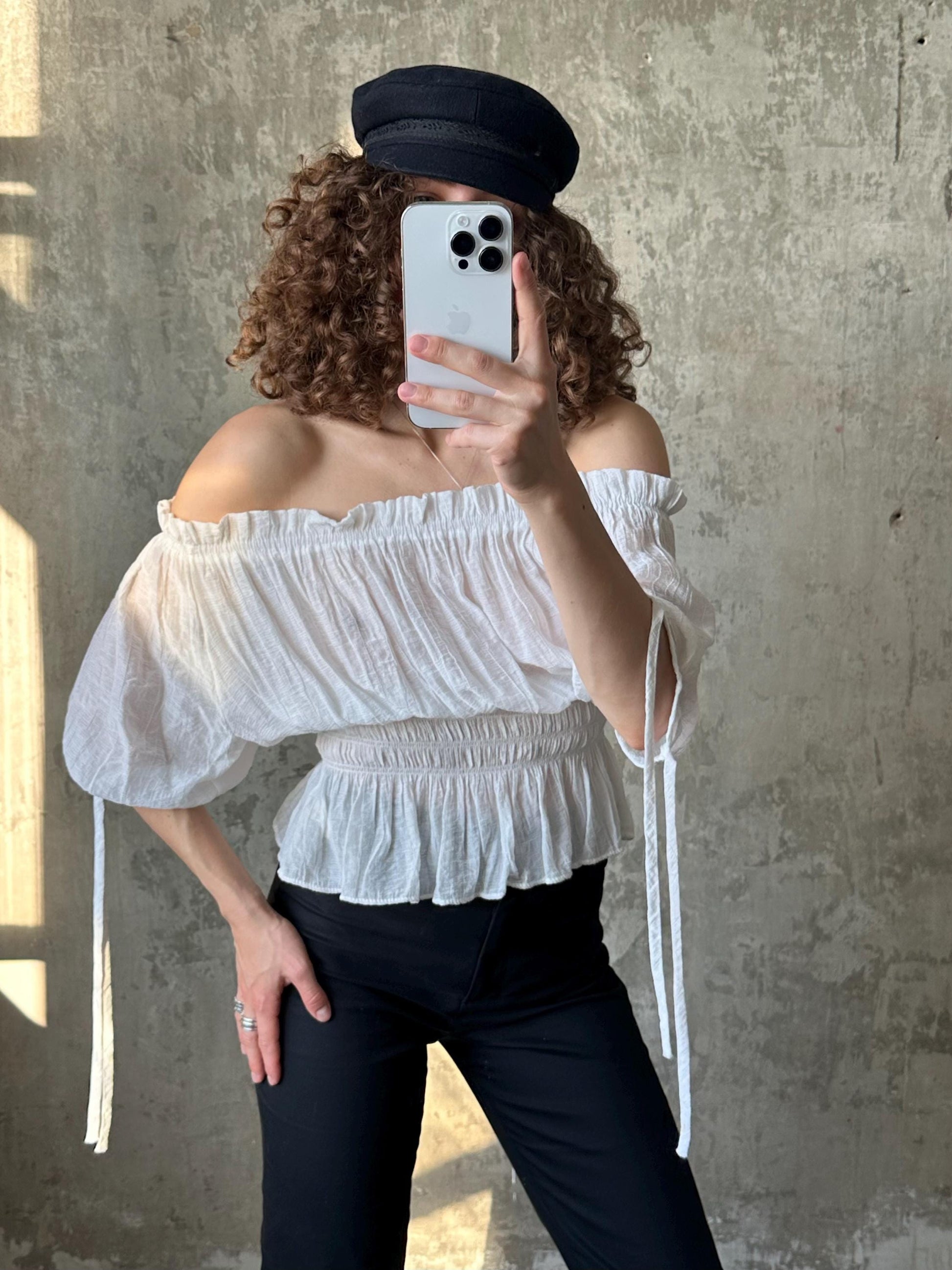 A person is taking a selfie with a white smartphone, wearing a white off-the-shoulder top and black pants.
