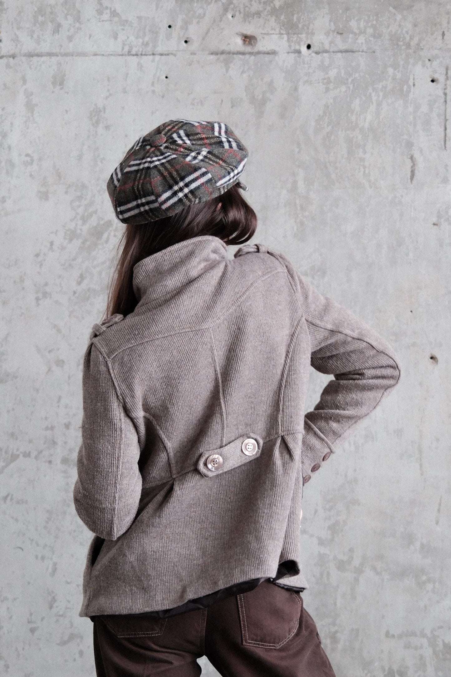 A person wearing a gray coat and plaid hat is standing against a concrete wall.