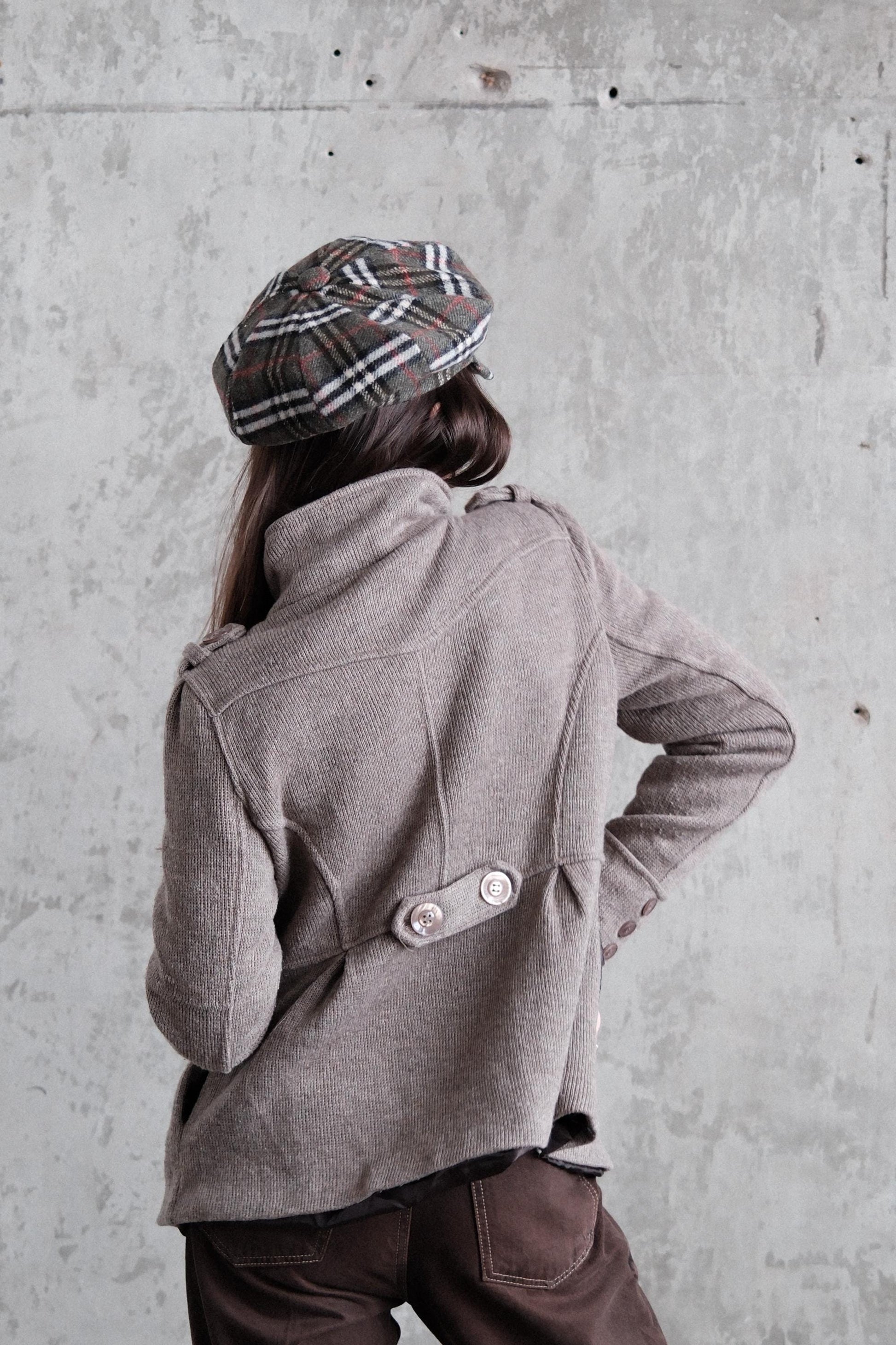 A person wearing a gray coat and plaid hat is standing against a concrete wall.