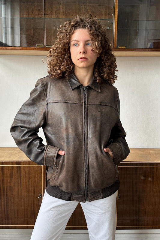 A woman with curly hair wearing a brown leather jacket stands in front of a wooden cabinet.