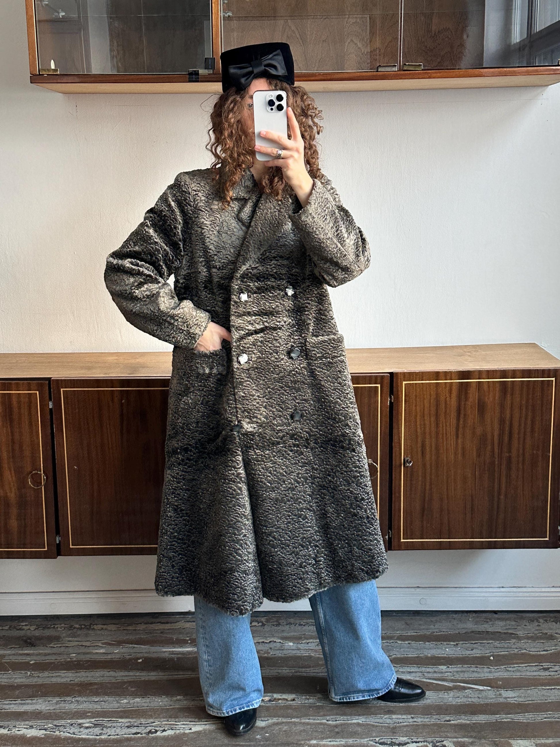 A person wearing a long gray coat and blue jeans is taking a selfie with a smartphone.