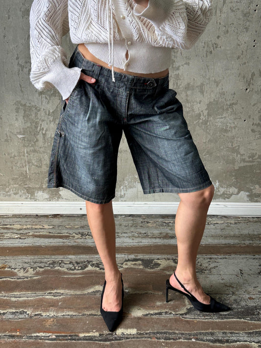 A person wearing a white sweater, blue jeans, and black heels stands in front of a concrete wall.