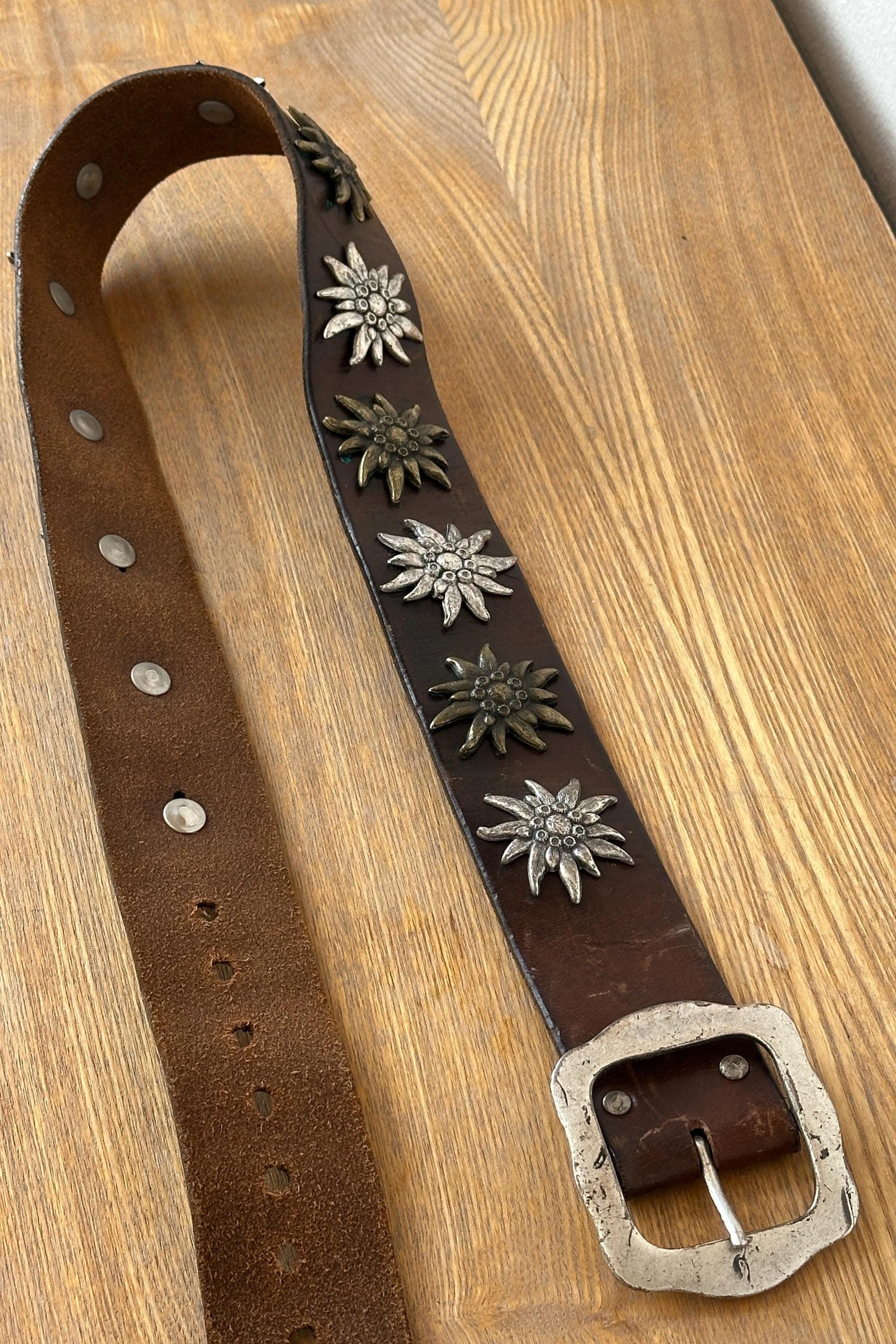 A brown leather belt with metal studs and a metal buckle is displayed on a wooden surface.