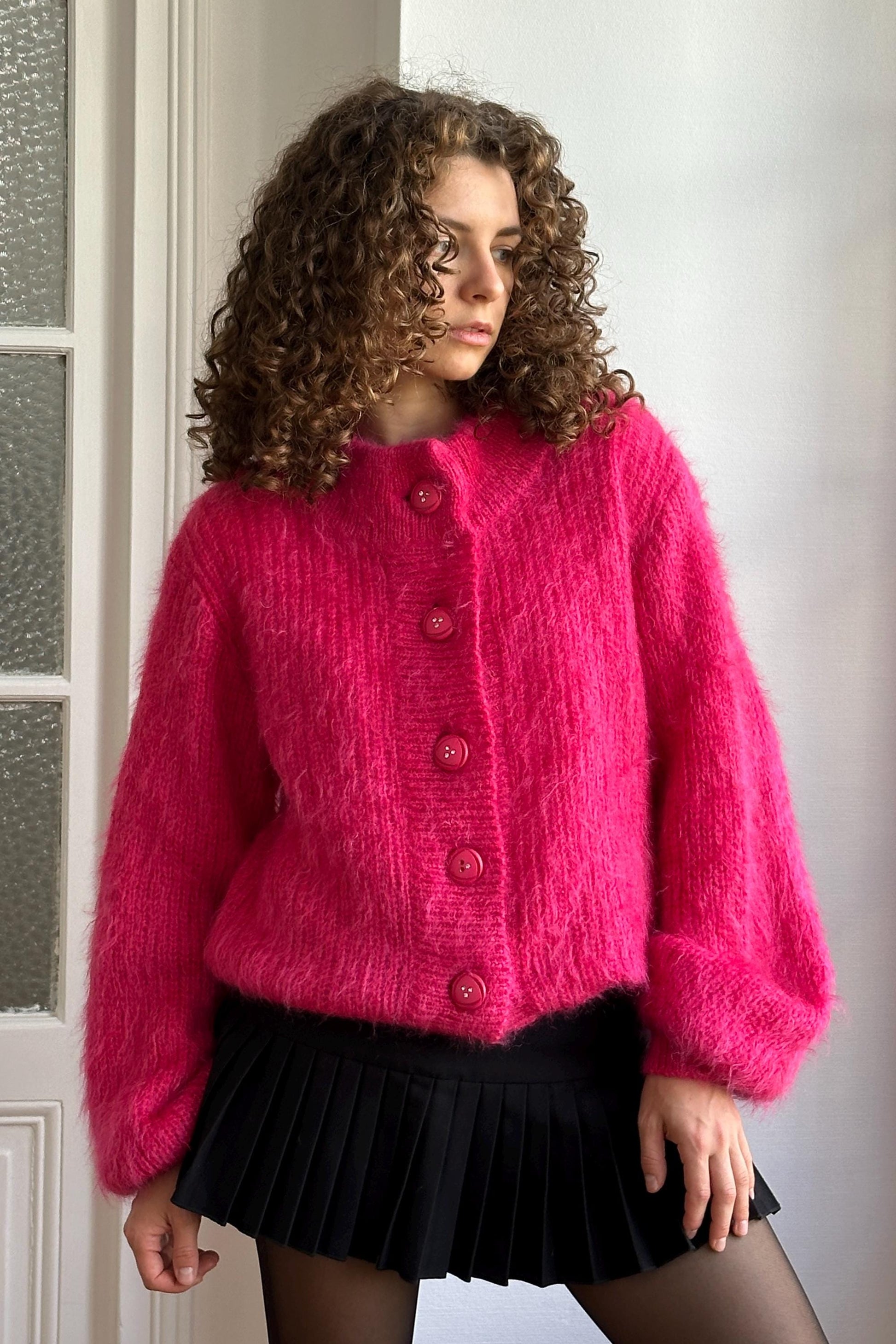 A woman with curly hair wearing a bright pink cardigan and a black pleated skirt stands in front of a white door.
