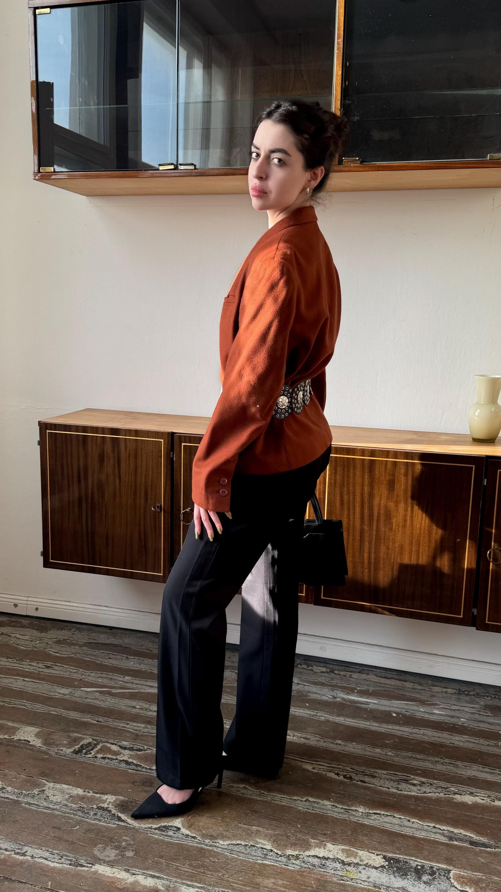 A woman in a red blazer stands in a room with wooden floors and cabinets, looking directly at the camera.