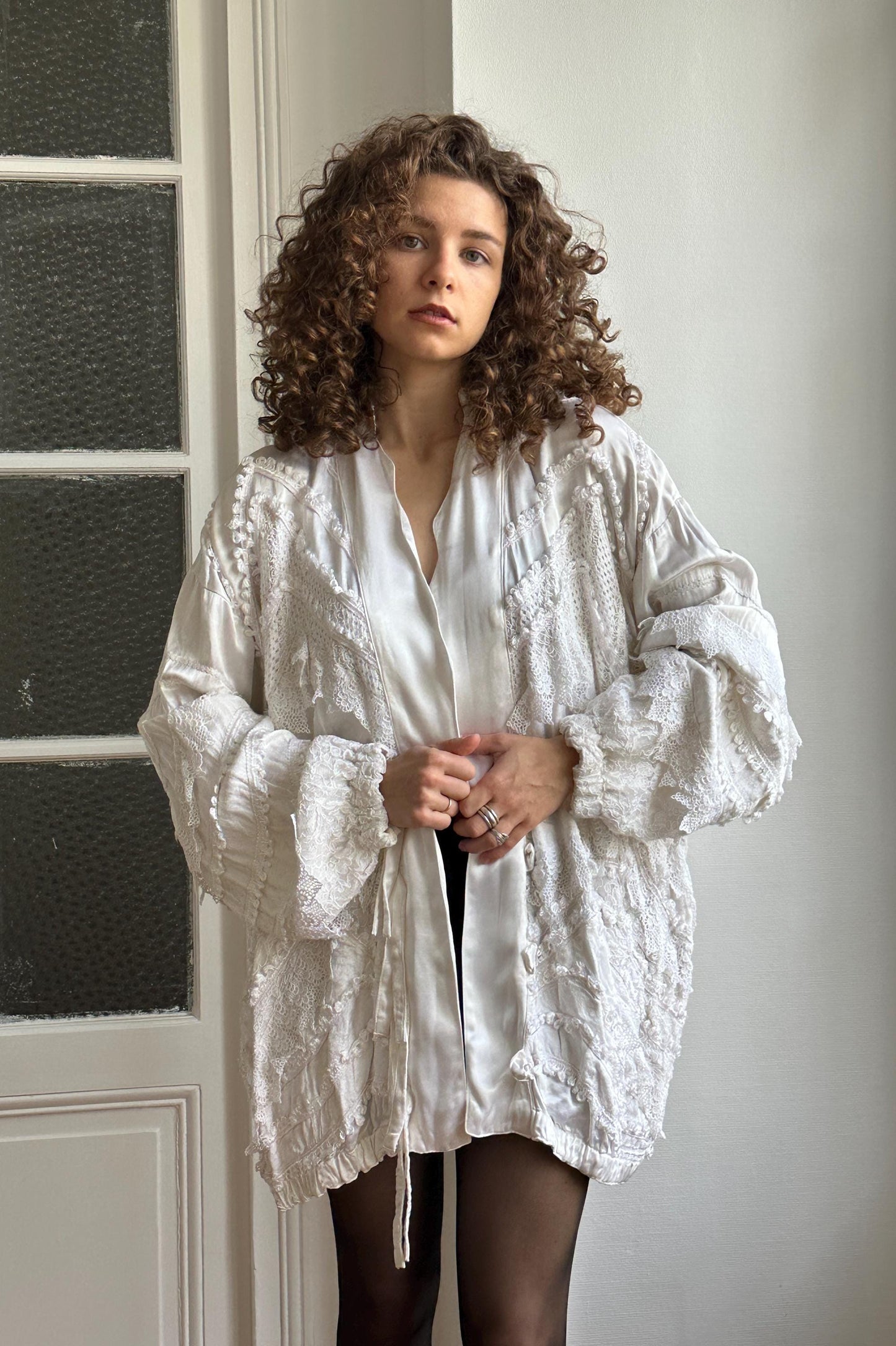 A young woman with curly hair wearing a white, lace-like jacket stands in front of a white wall, looking directly at the camera.