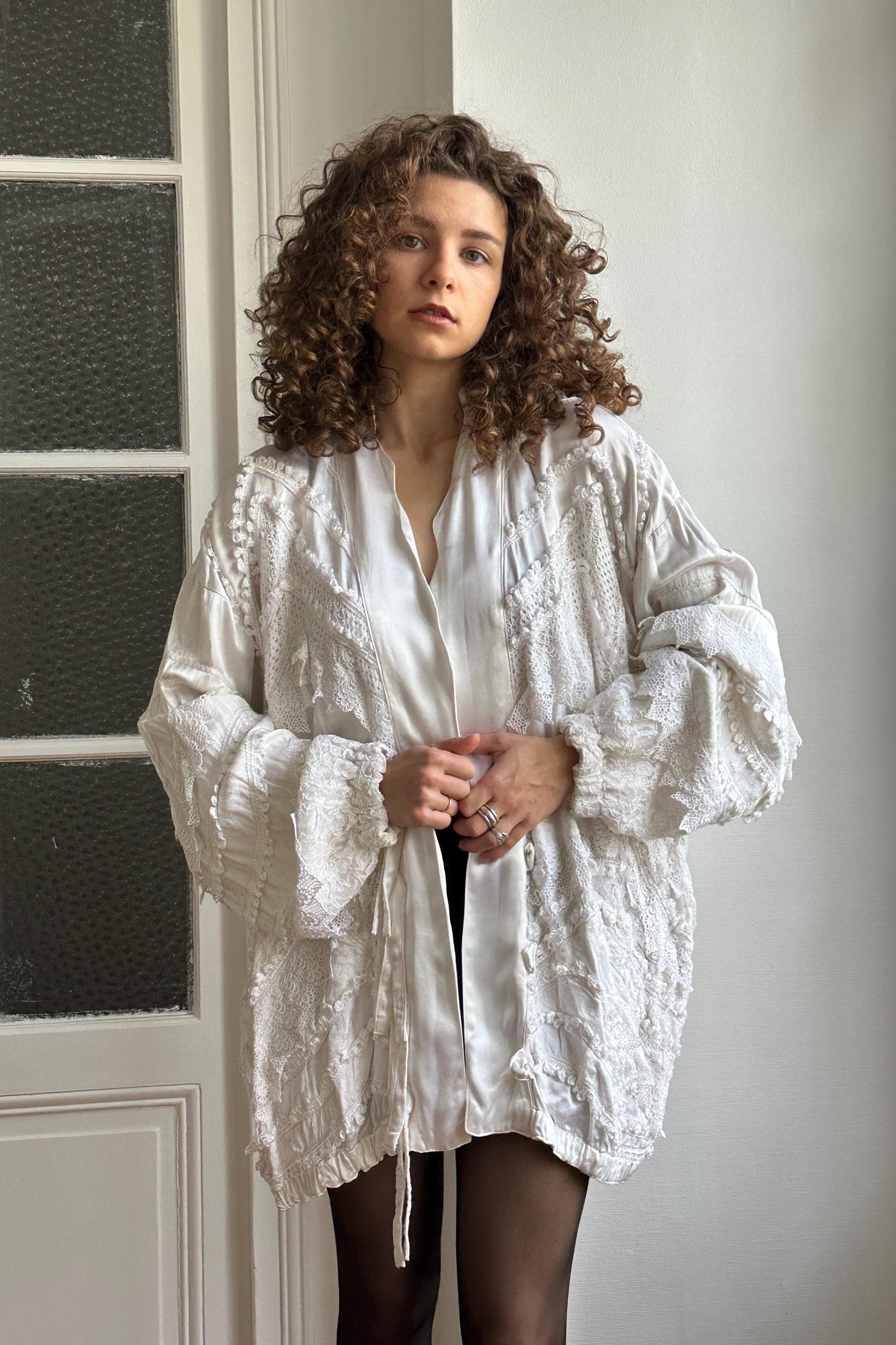A young woman with curly hair wearing a white, lace-like jacket stands in front of a white wall, looking directly at the camera.