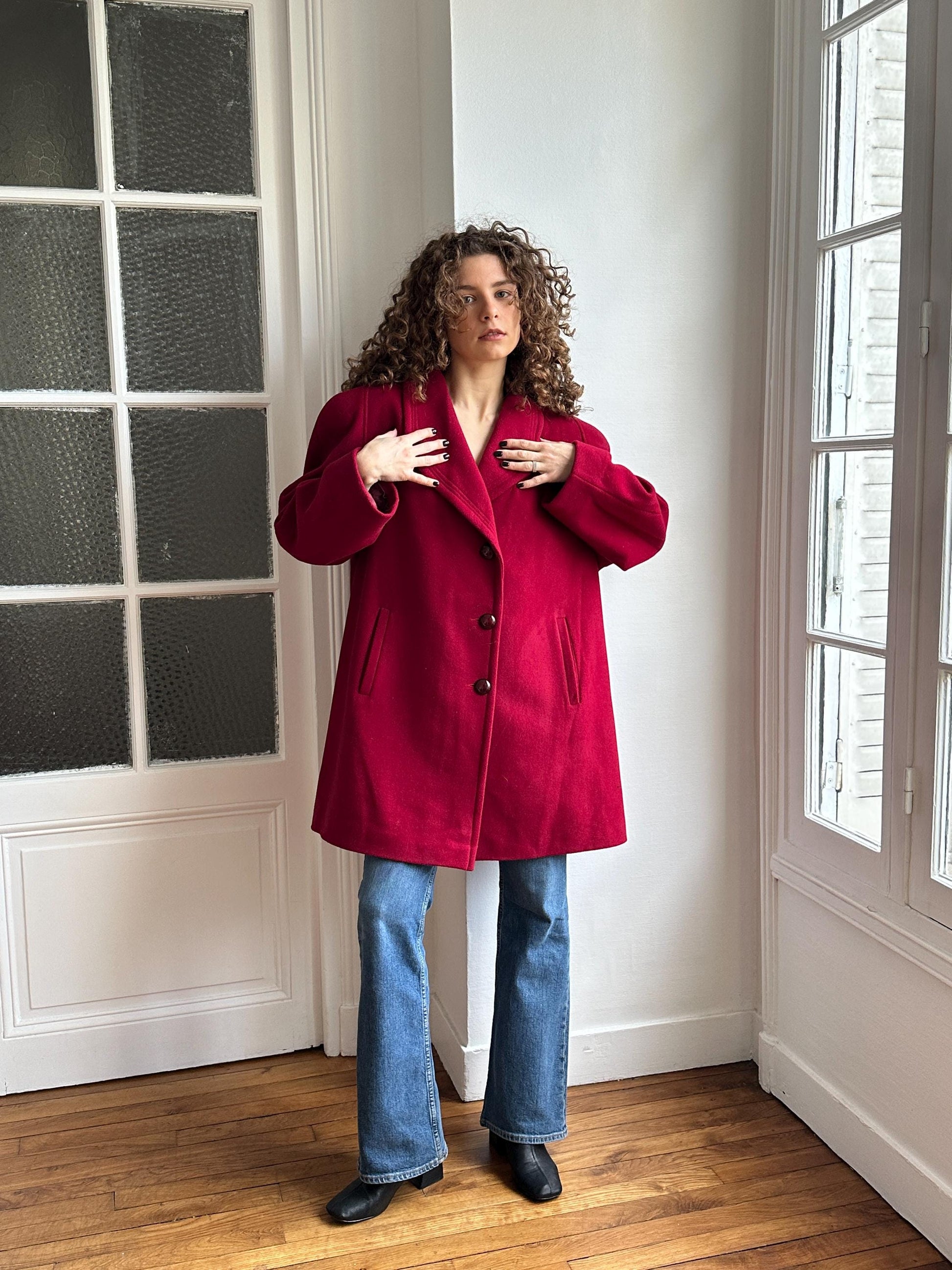 A person in a red coat stands in a room with a window, their hands raised to their head.