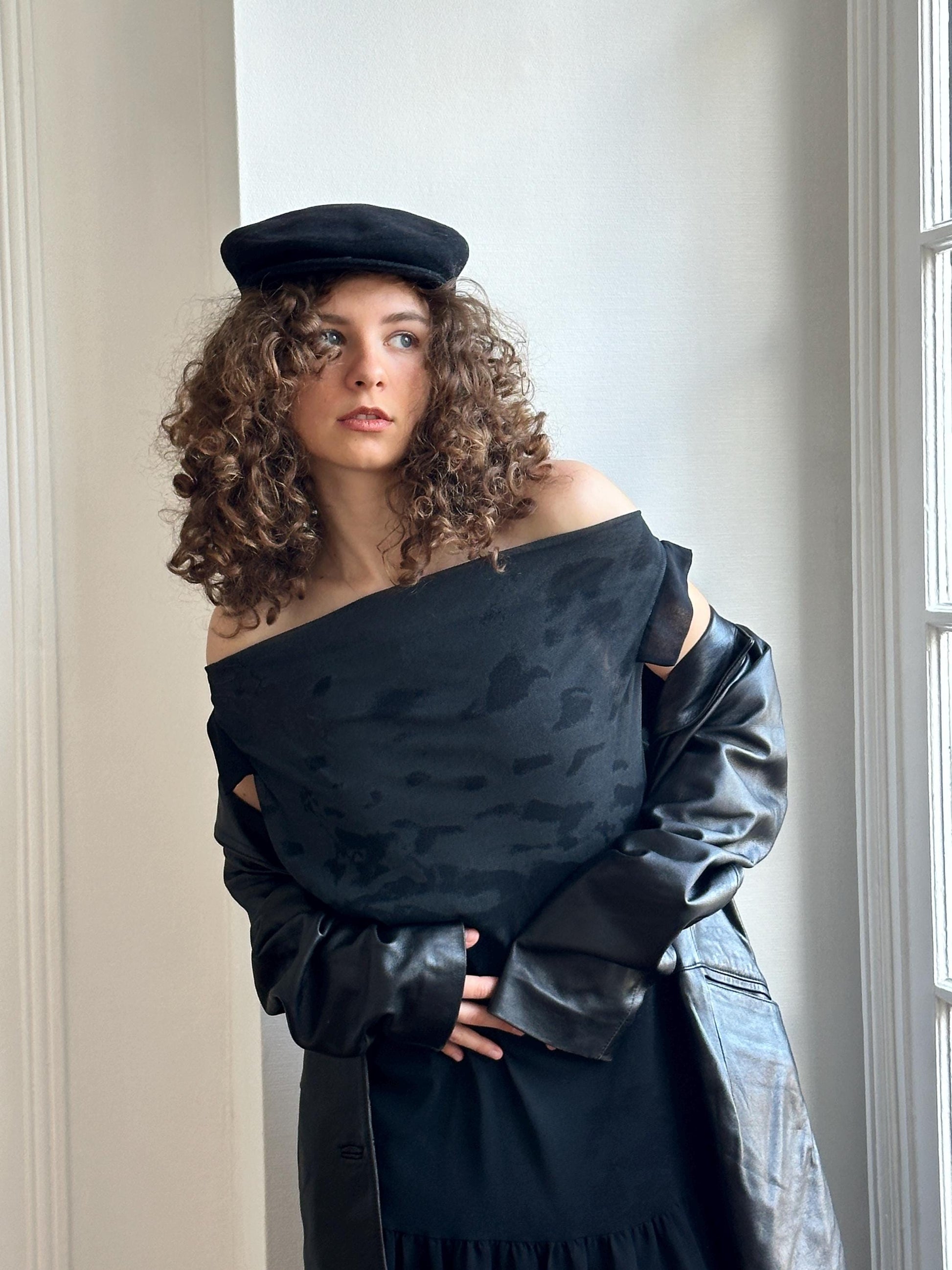 A woman with curly hair wearing a black off-the-shoulder top and a black hat stands in front of a white wall, with a leather jacket draped over her shoulders.