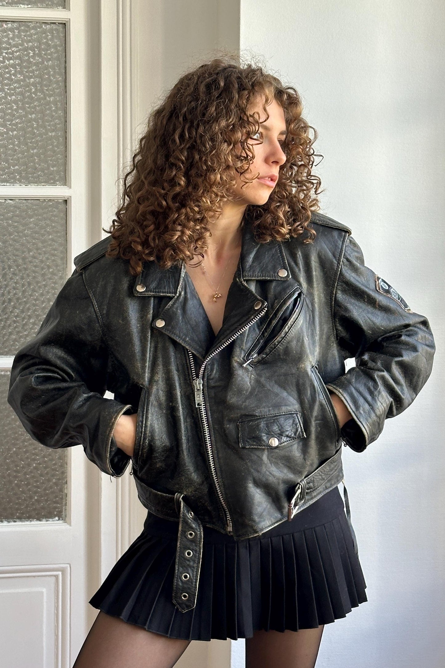 A woman with curly hair wearing a black leather jacket and a black pleated skirt stands in front of a white door.